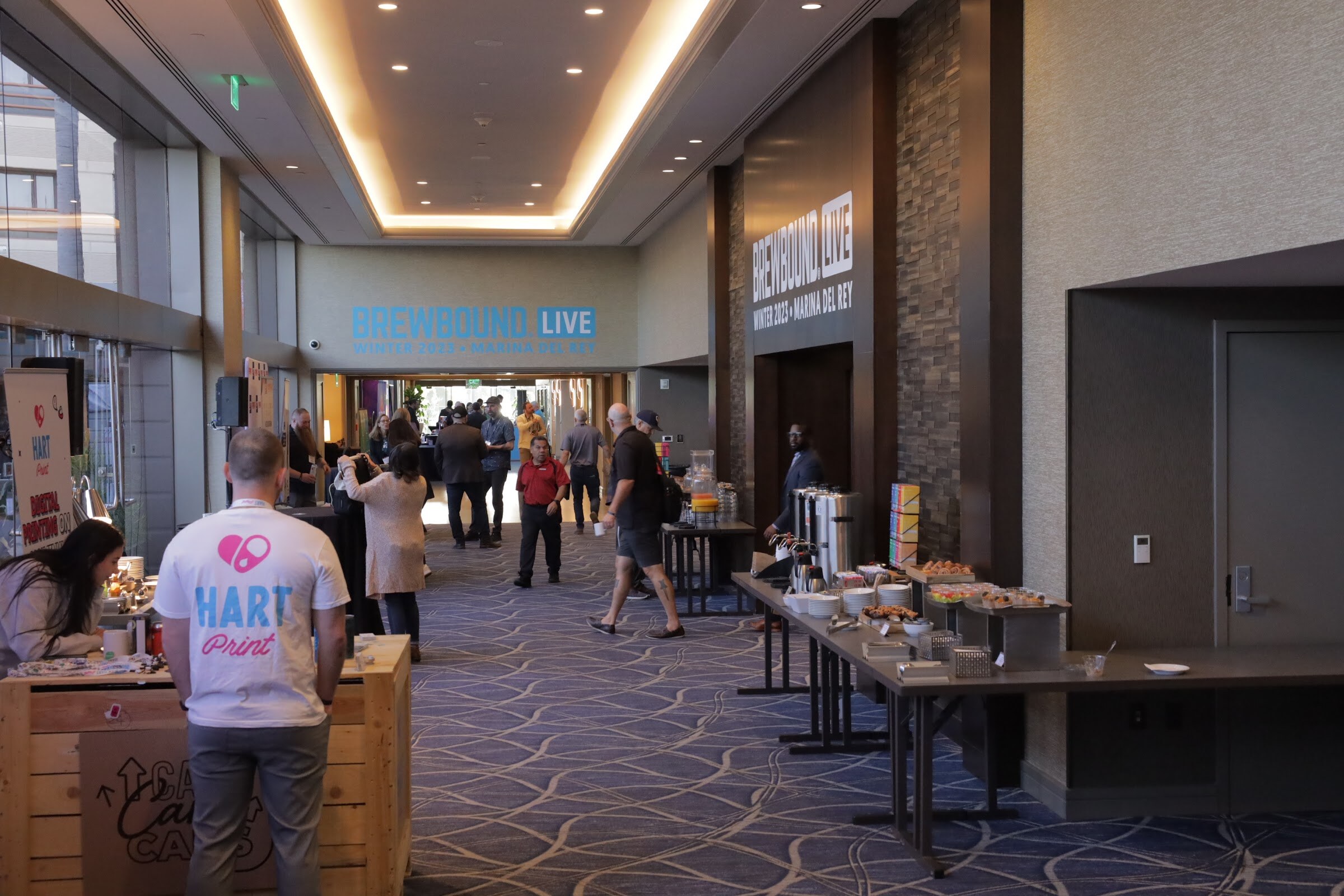 A long carpeted hallway with a table of coffee and snacks and attendees milling along it, with a Brewbound Live sign at the end.
