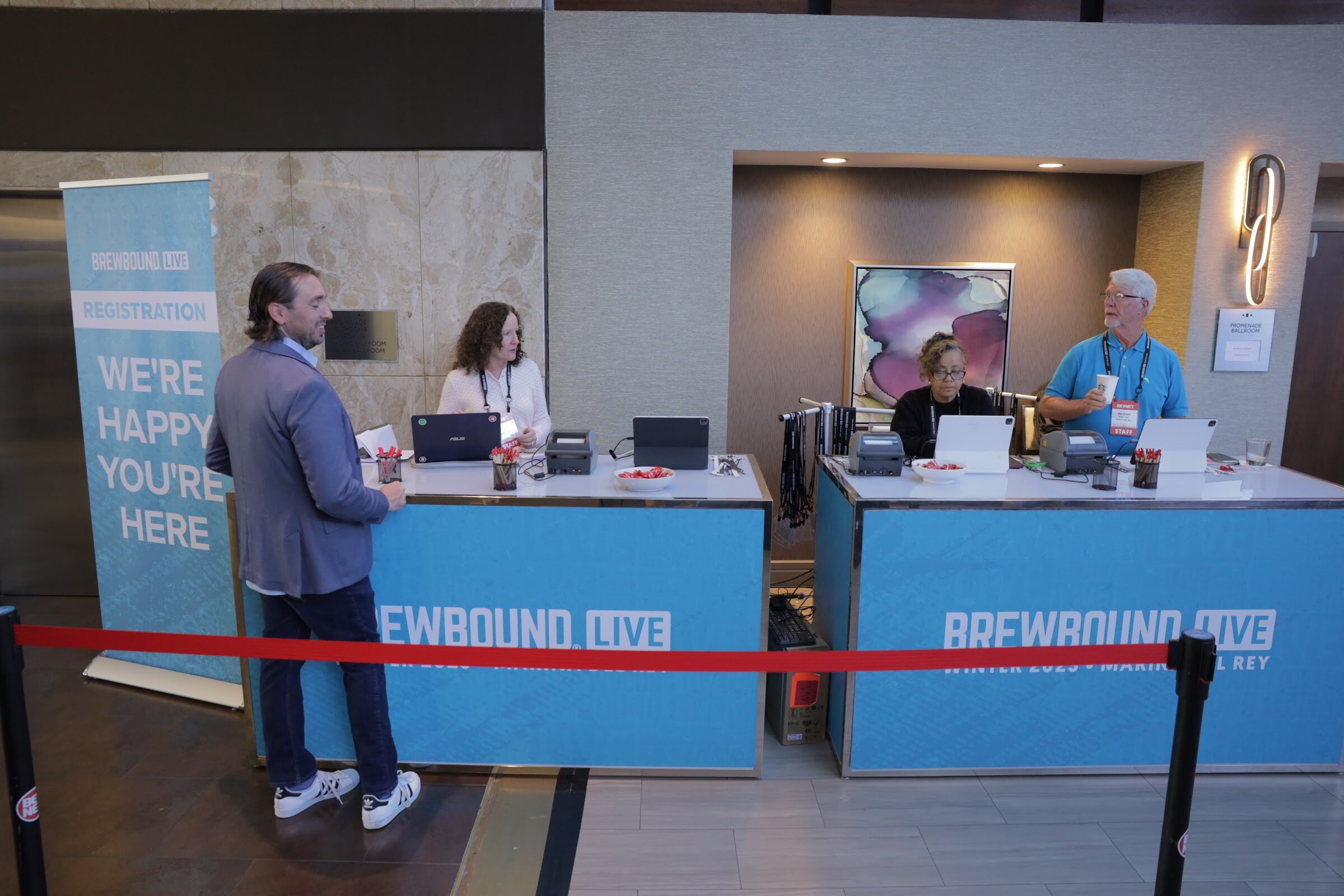 Four people stand and sit at two blue Brewbound Live registration desks with laptops and a red rope line.