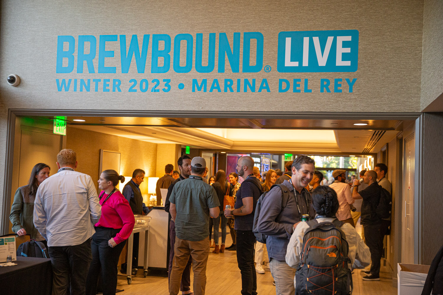 A crowd of attendees talks in a doorway beneath a teal Brewbound Live Winter 2023 Marina del Rey sign.