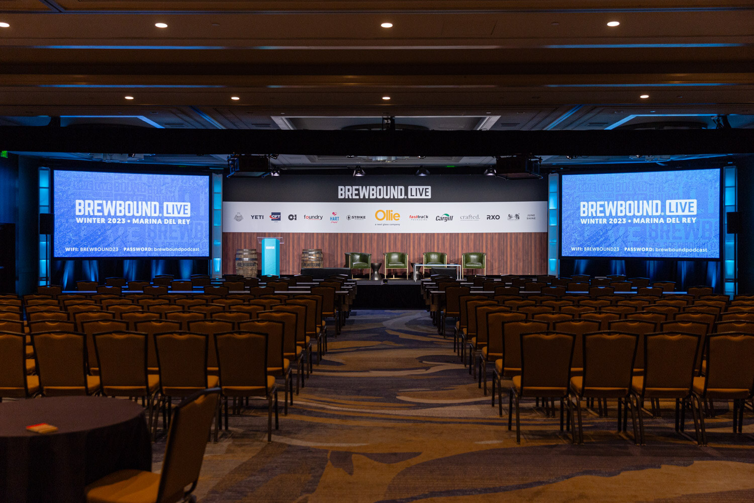 A ballroom set with rows of chairs facing a stage with two large blue screens reading Brewbound Live Winter 2023 Marina del Rey.