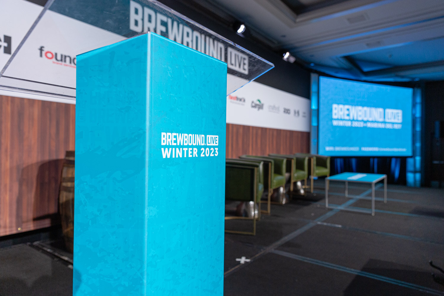 A teal lectern marked Brewbound Live Winter 2023 in front of a stage with green armchairs and a screen.