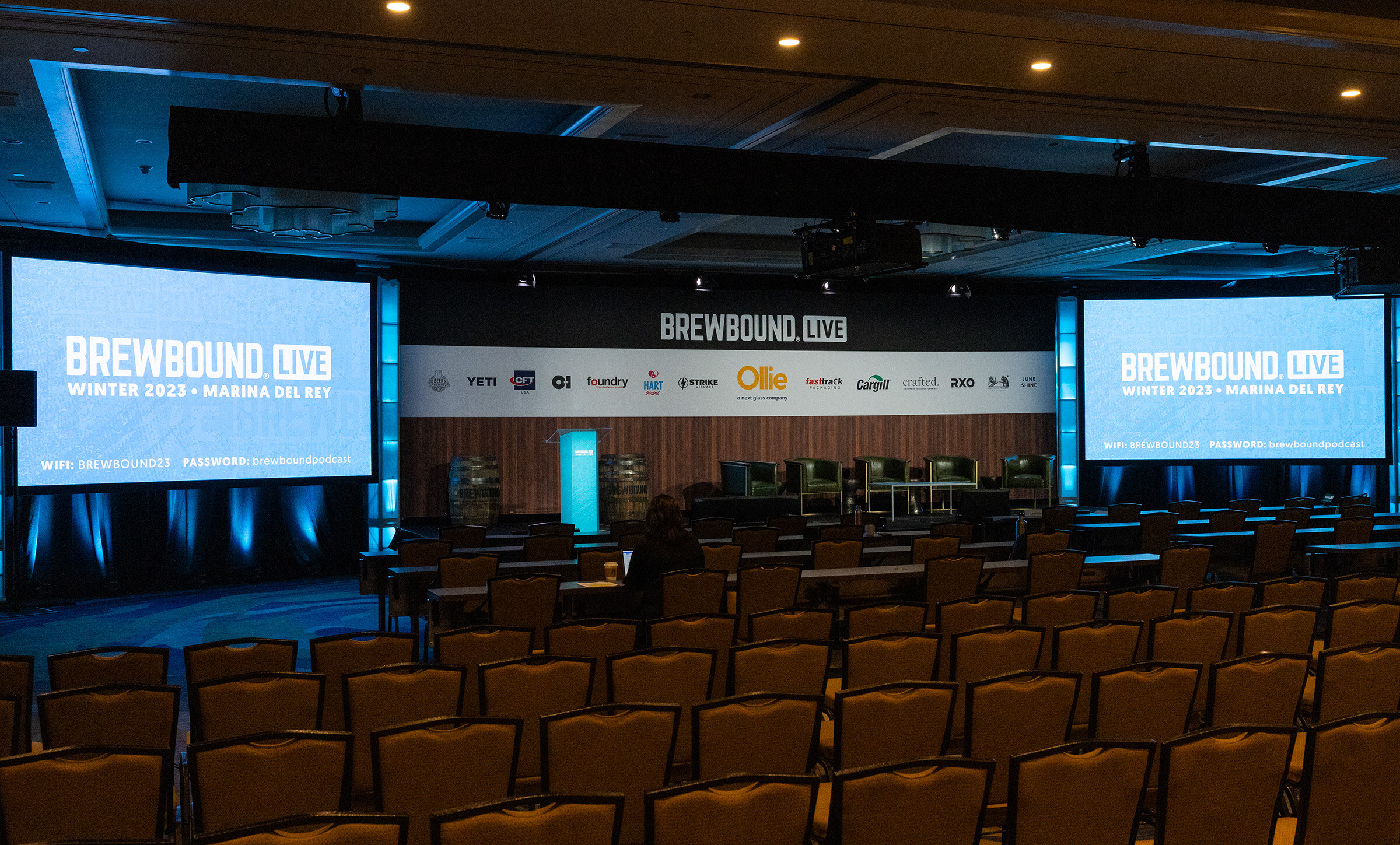 A dim ballroom with rows of empty chairs facing a stage and two large screens reading Brewbound Live Winter 2023 Marina del Rey.