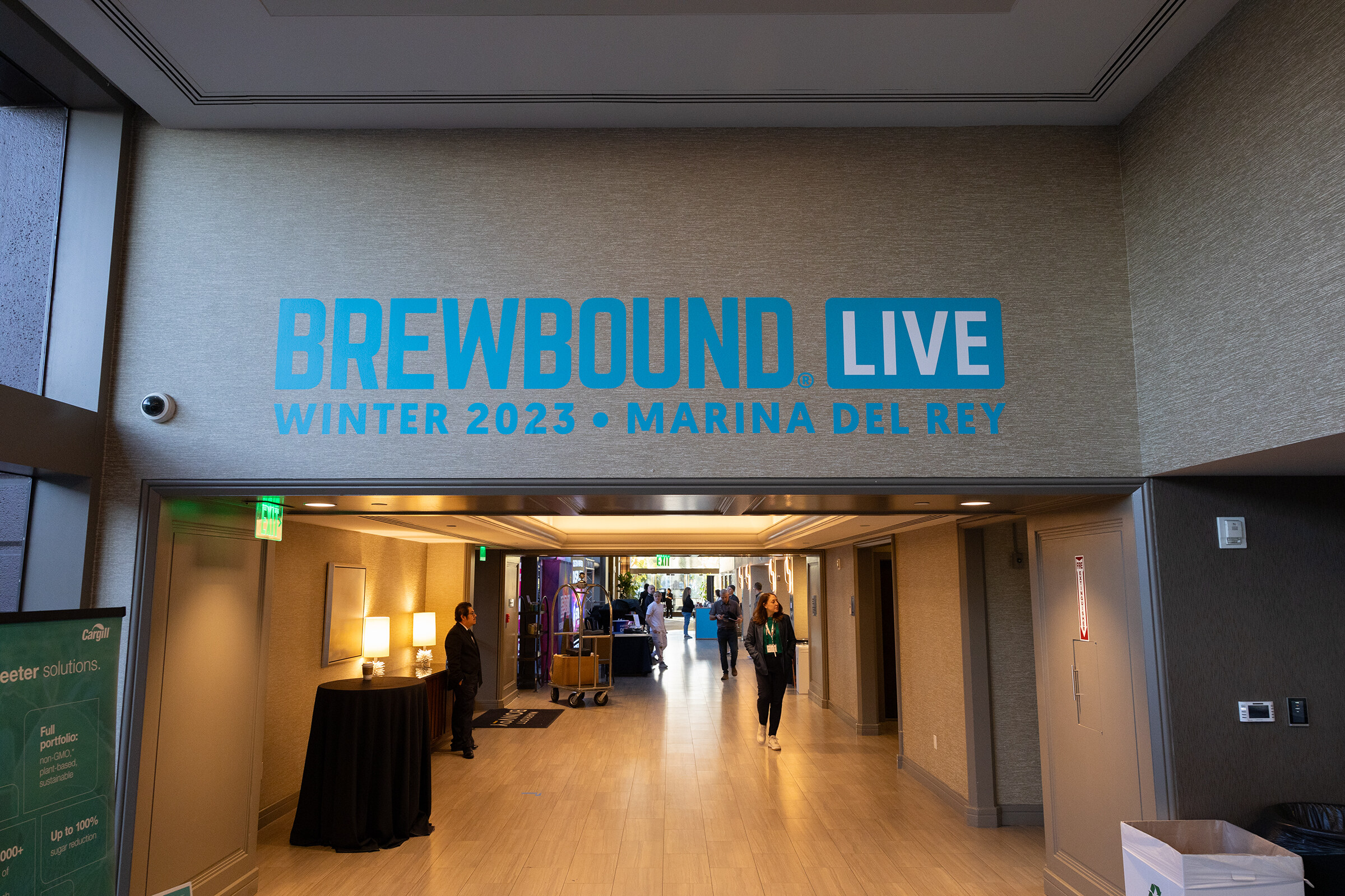 A beige hallway with a teal Brewbound Live Winter 2023 Marina del Rey sign above a corridor with a few attendees.