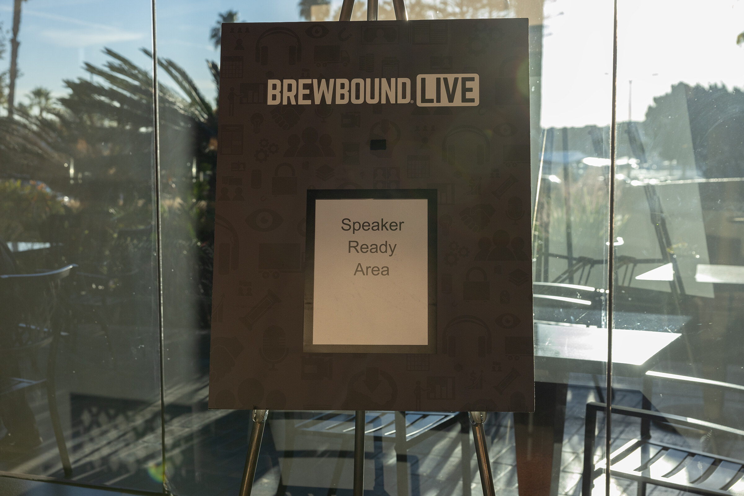 An easel sign with a Brewbound Live logo and the text Speaker Ready Area, with palm trees visible through glass behind it.