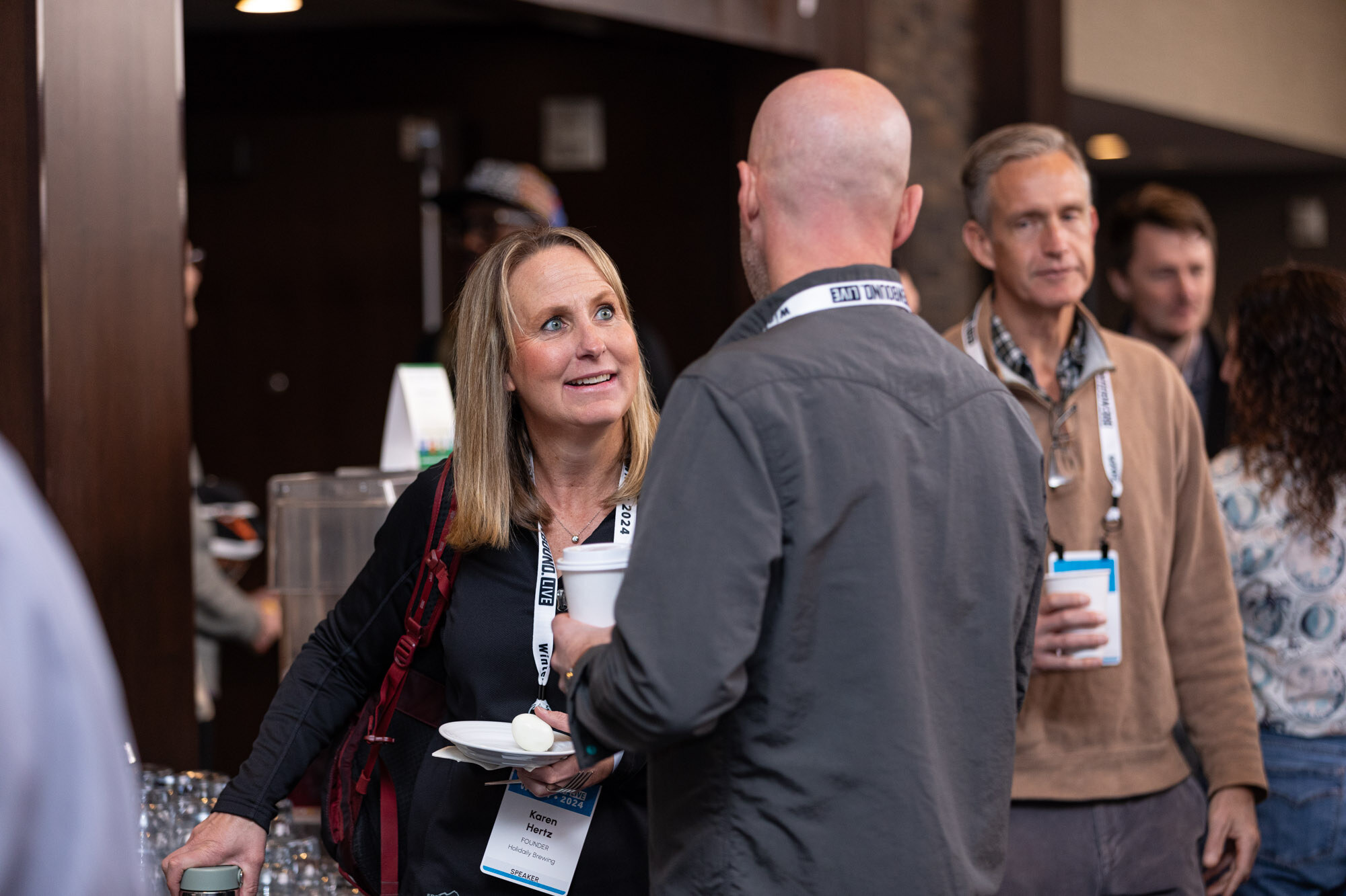 A smiling woman with a plate talks with a bald man and another man near the entrance.