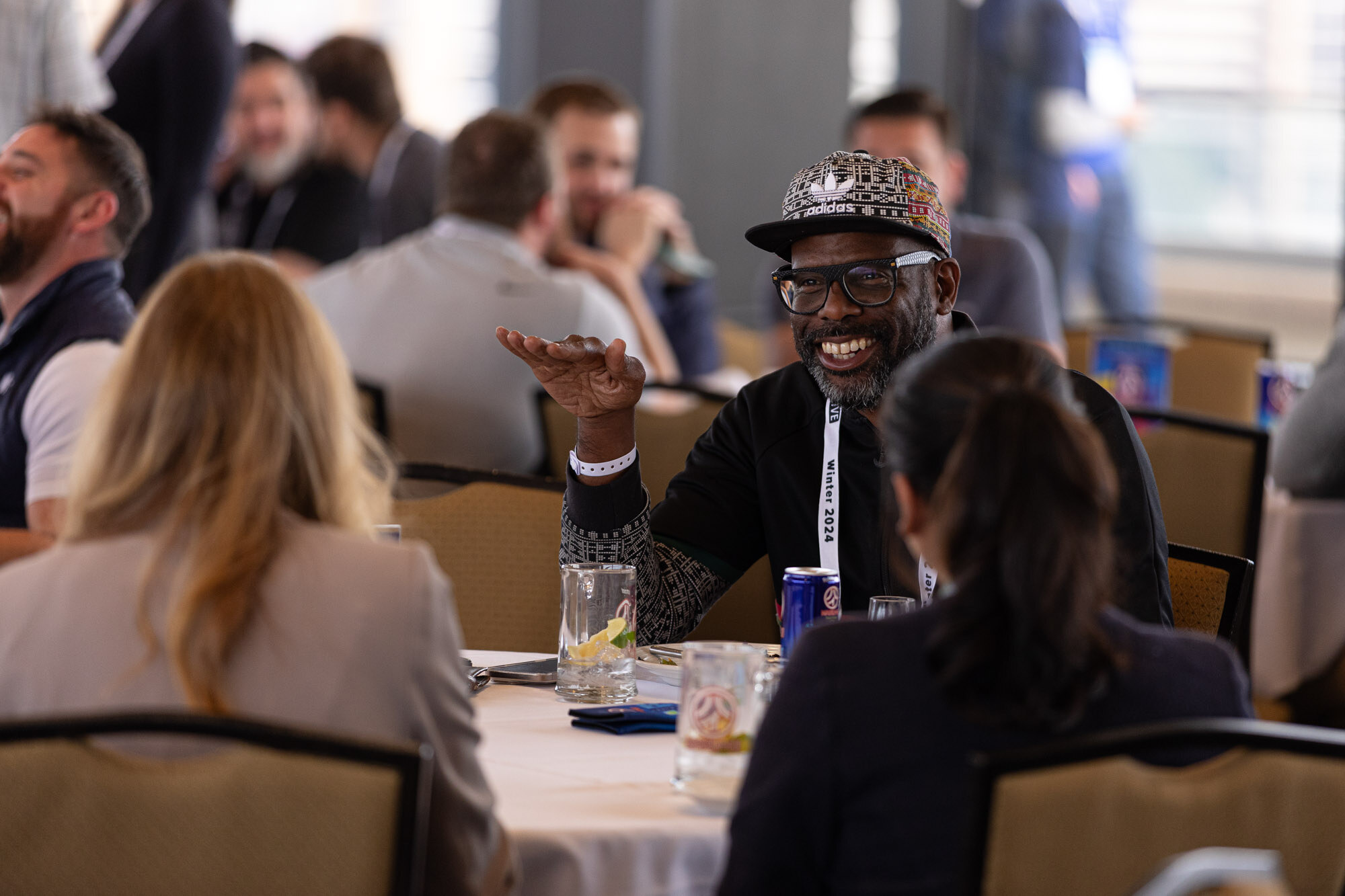 A man in a patterned cap and glasses smiles and gestures while seated at a round table.