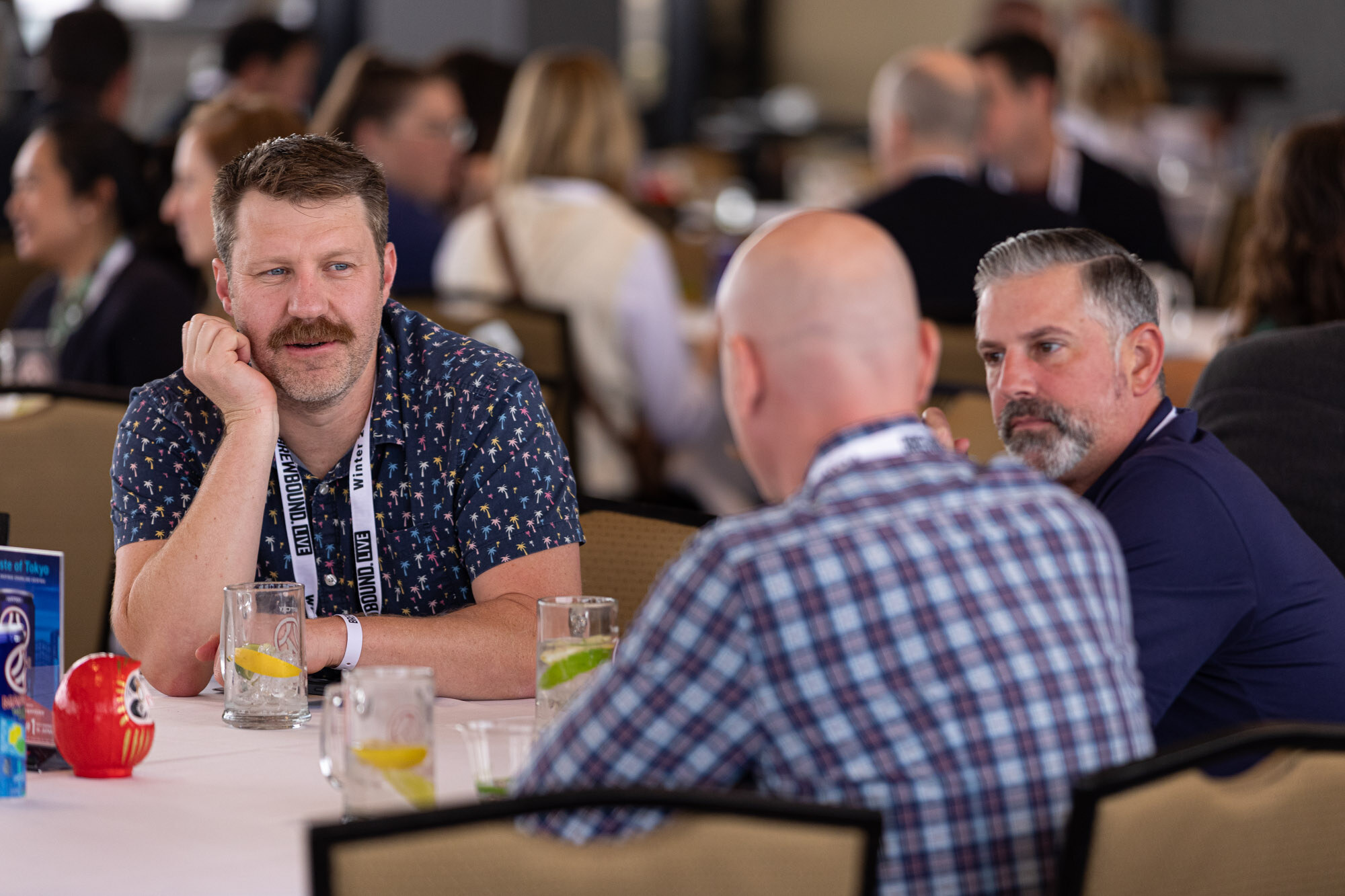 A man with a mustache rests his chin on his hand next to two other men at a table.