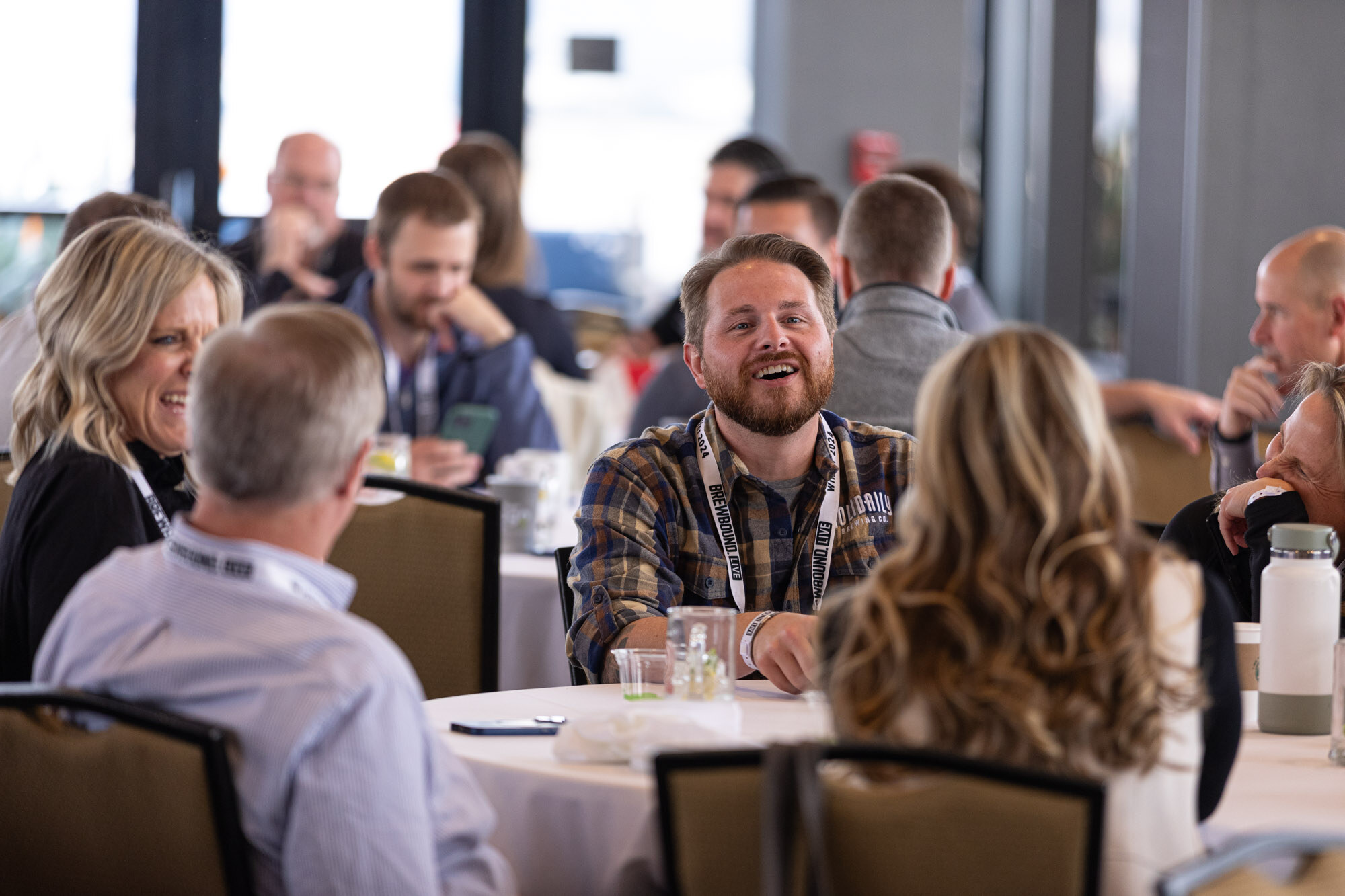A man in a plaid shirt laughs at a crowded round table with other attendees.