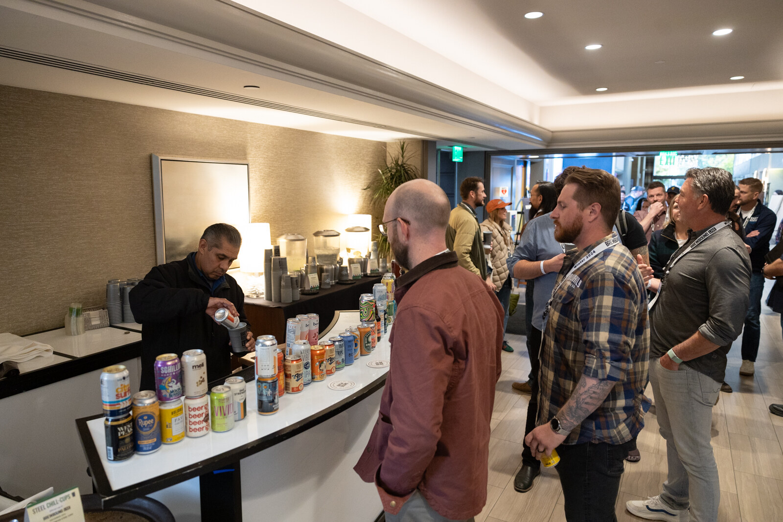 A bartender pours from cans at a curved white bar while attendees with badges stand nearby.