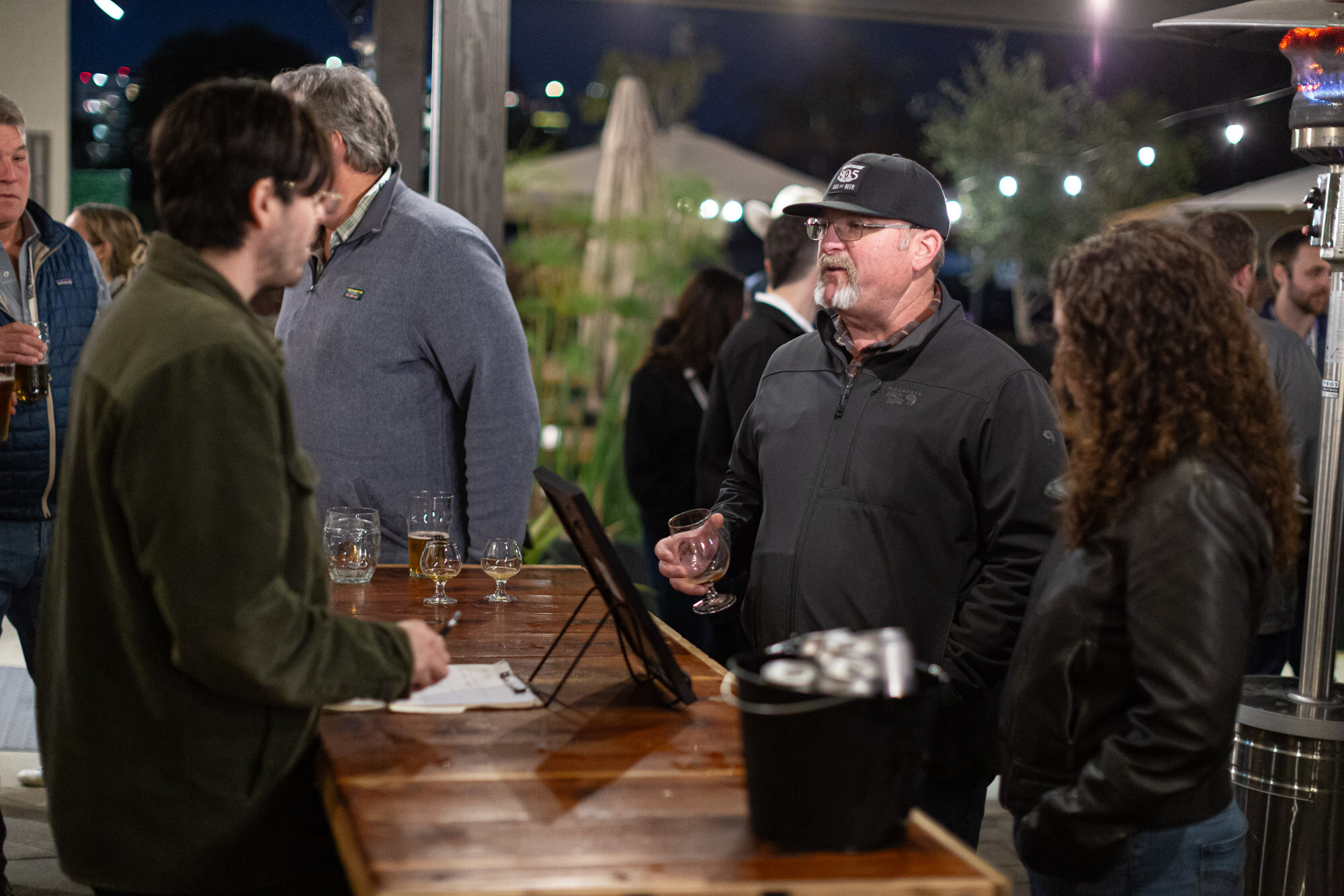 A group of attendees stands at a wooden table with glasses at night.