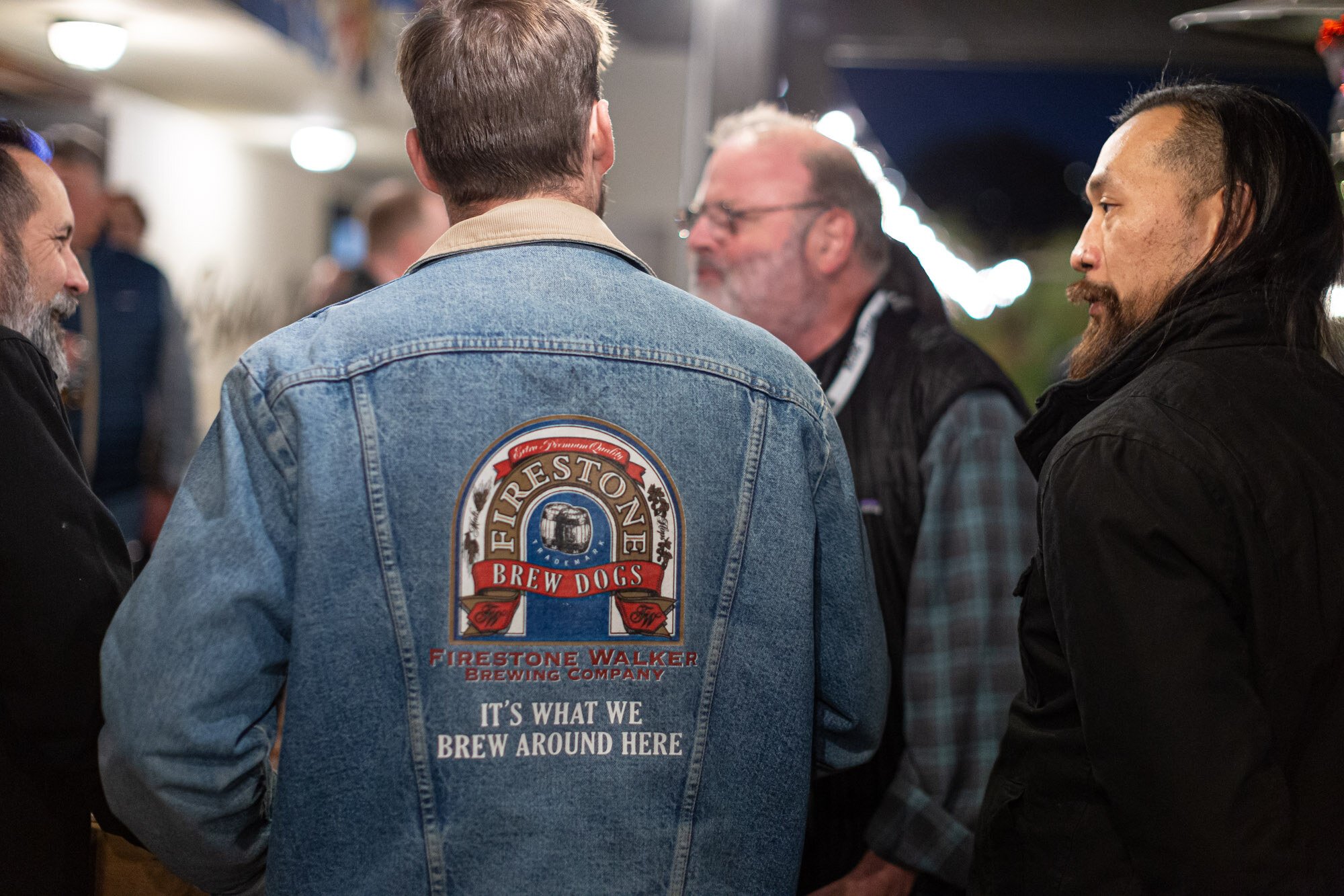 The back of a denim jacket with a Firestone Brew Dogs patch as two other men look on.