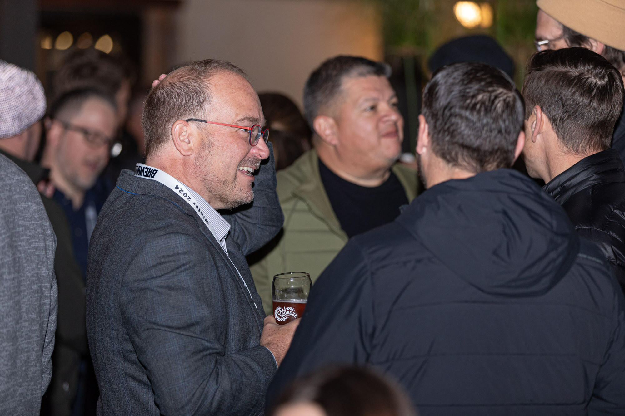 A smiling man in a gray blazer and glasses talks with a group in a crowded room.