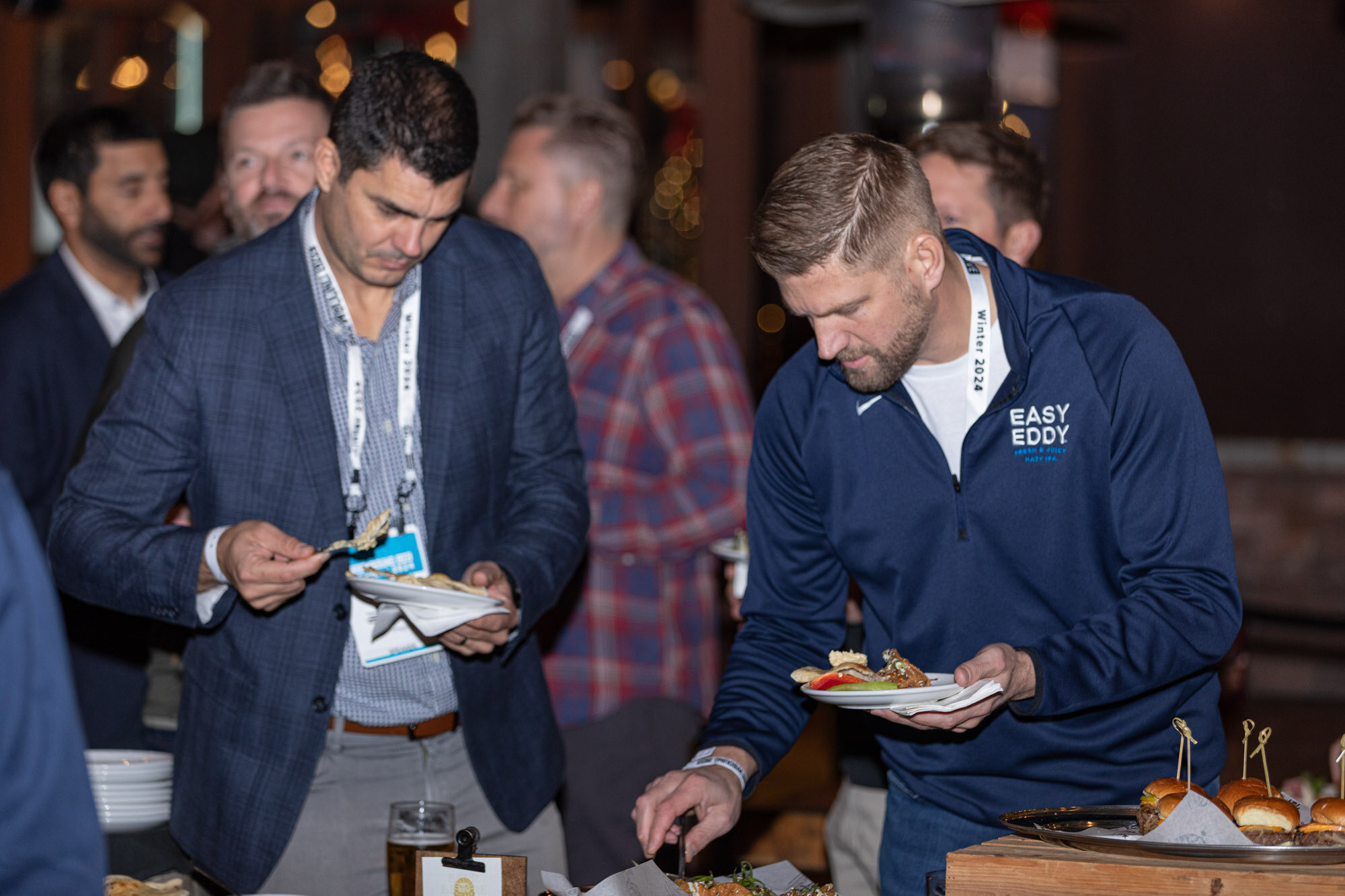 Two men in blazers and jackets hold plates at a food table.