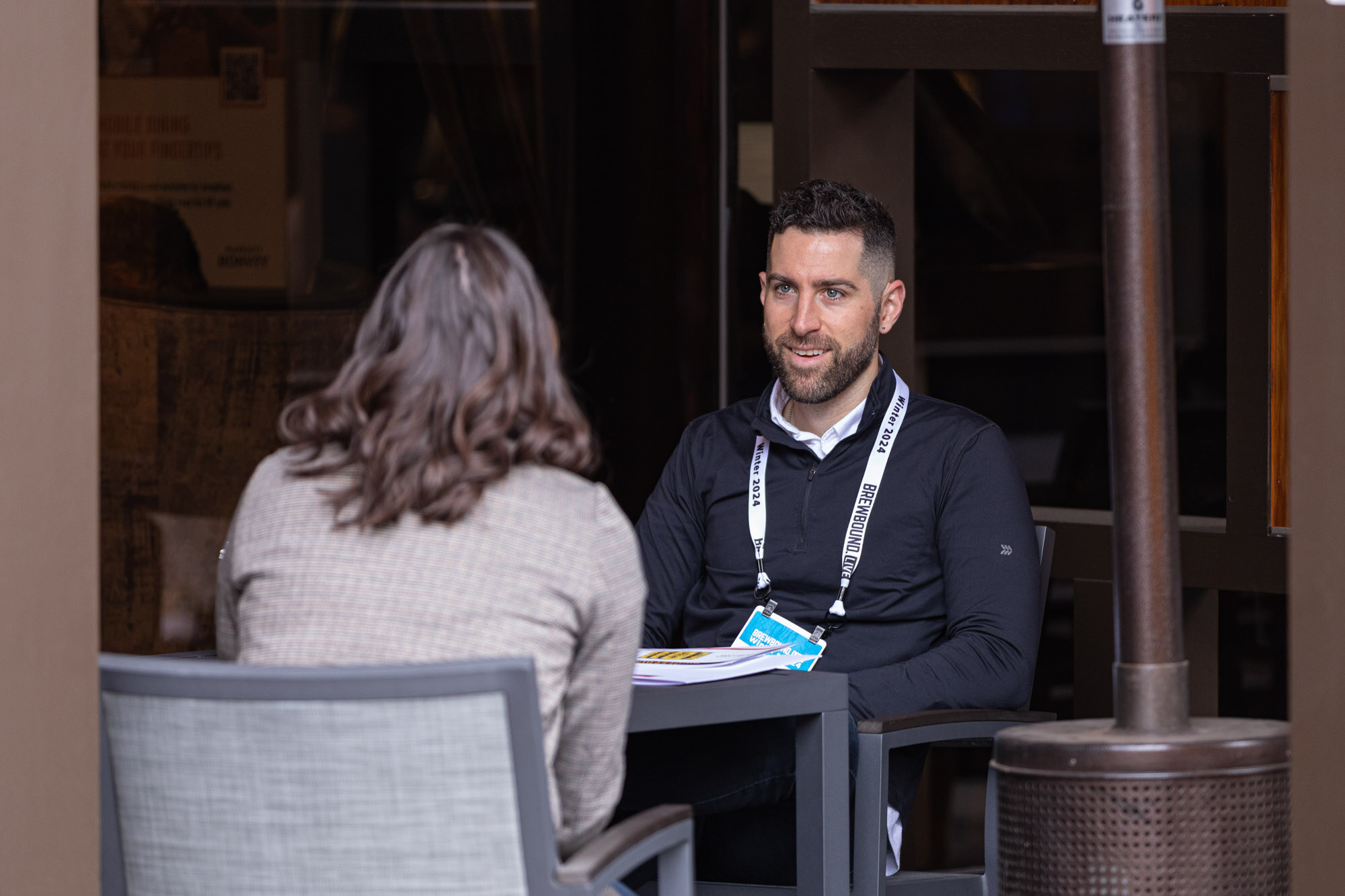 A smiling man with a lanyard sits at a table across from a woman seen from behind.