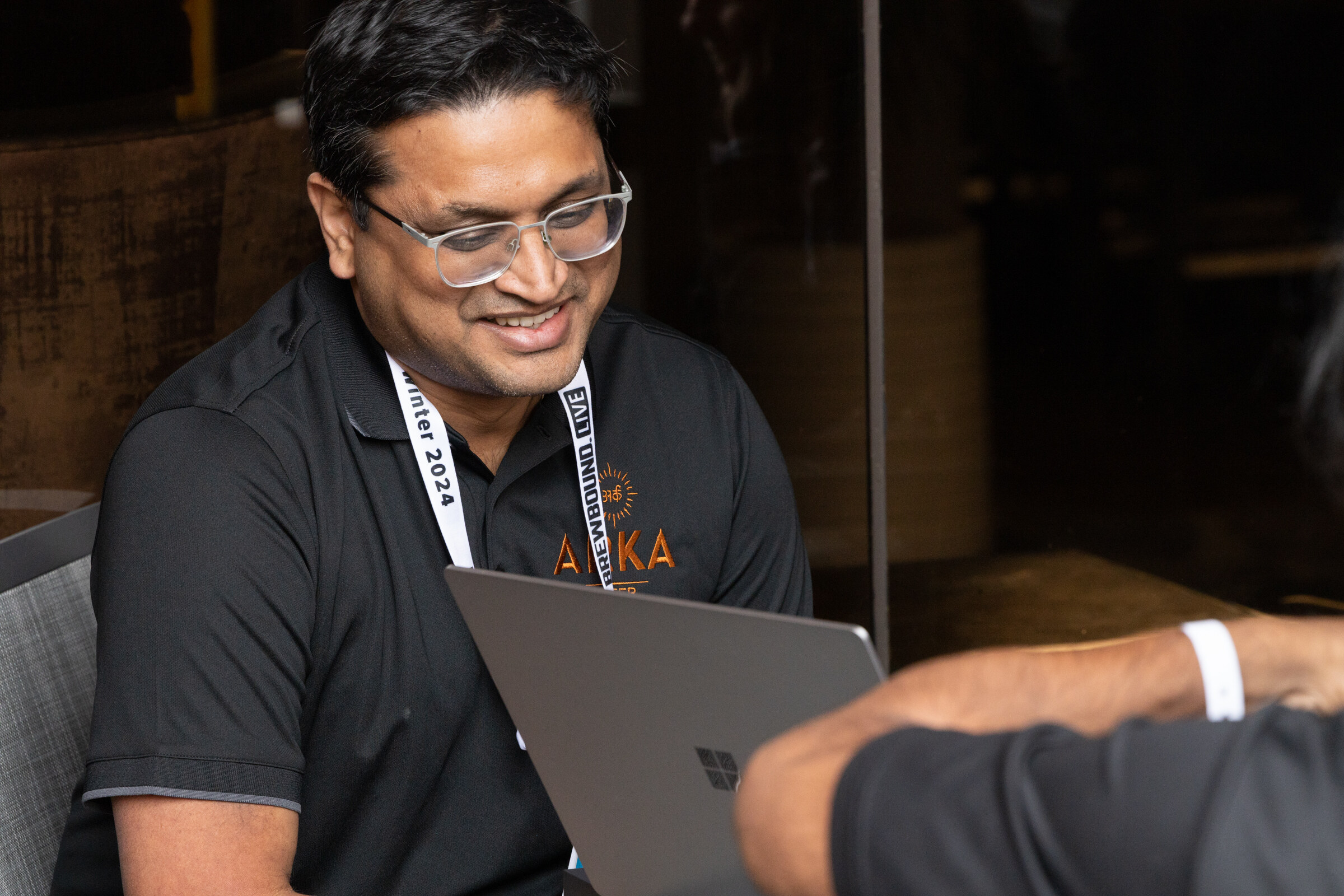A man in glasses and a black Arka shirt smiles over a laptop at an outdoor table.