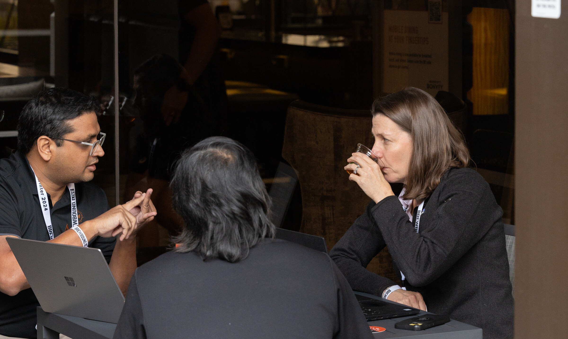 A woman sips from a small glass at a table while two men sit with a laptop.