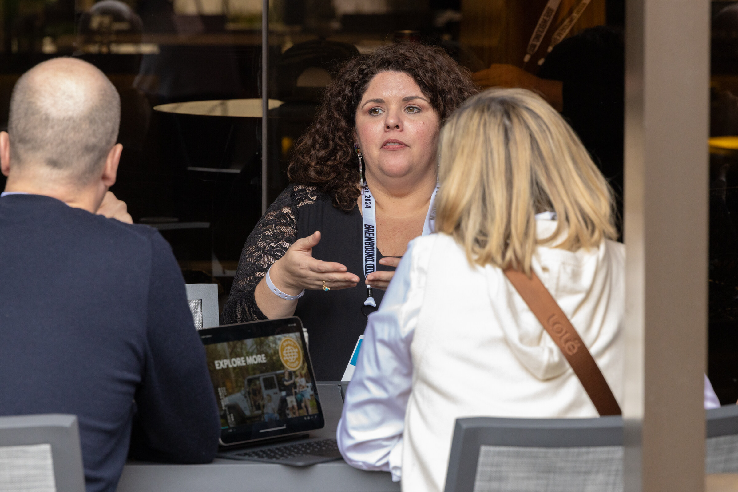 A woman with curly dark hair gestures while talking to two people at a table.