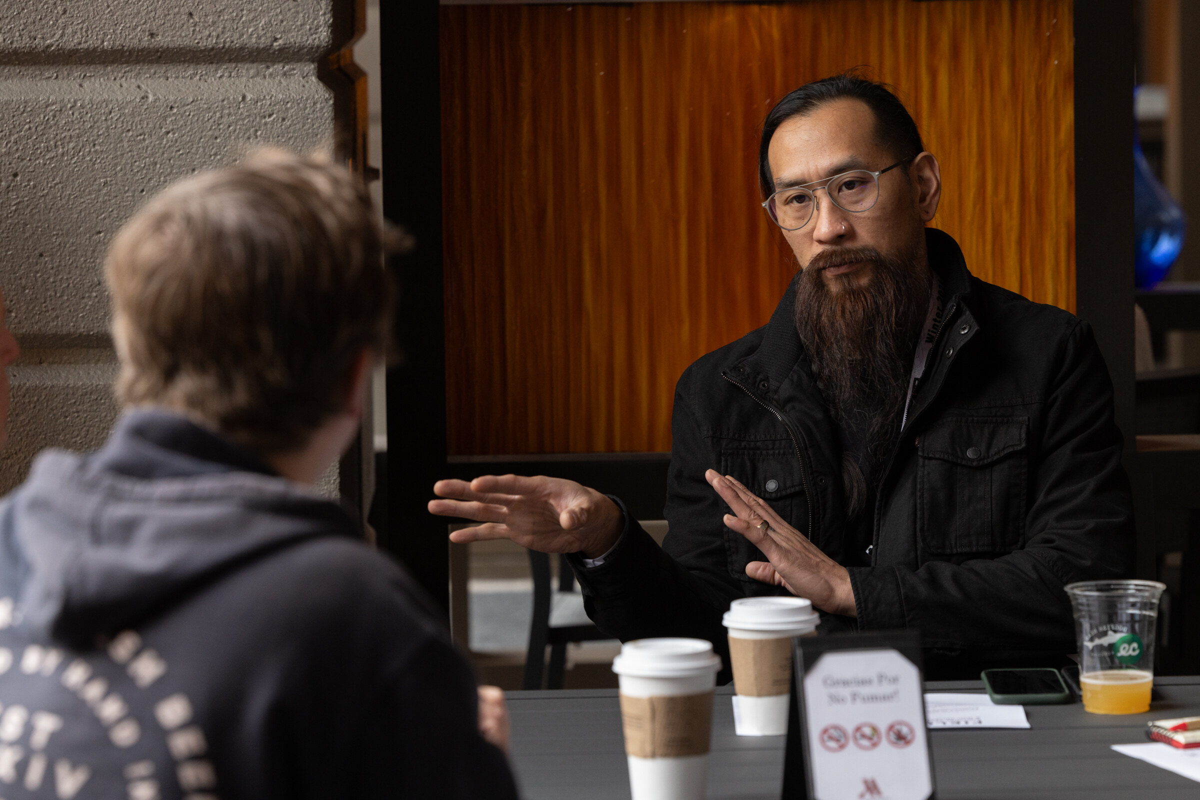 A man with a long beard and glasses holds a hand out while talking to a man in a hoodie.