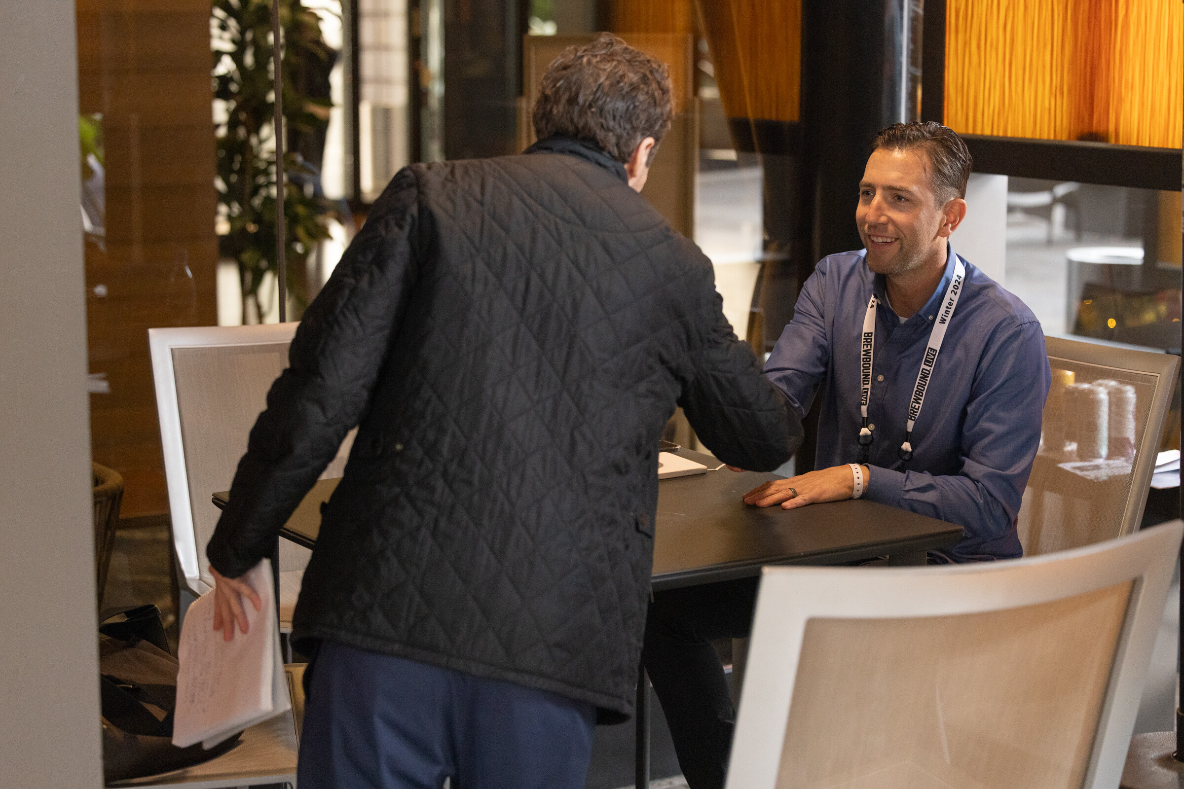 A man in a quilted jacket shakes hands with a seated man in a blue shirt.