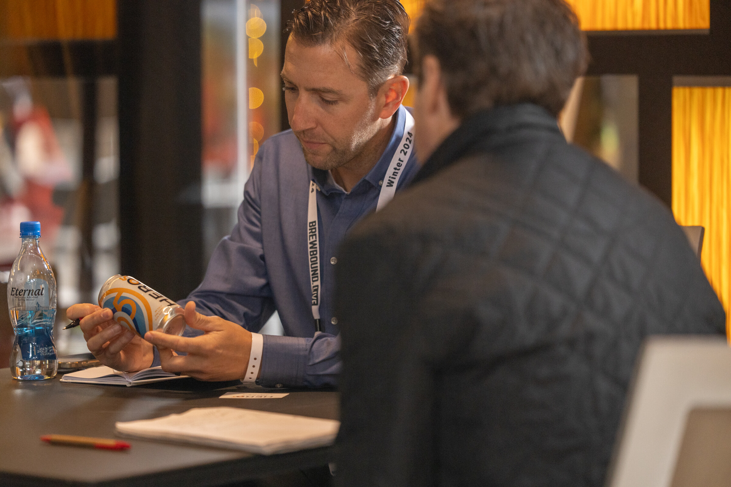 A man in a blue shirt holds a can while a man in a quilted jacket sits across the table.