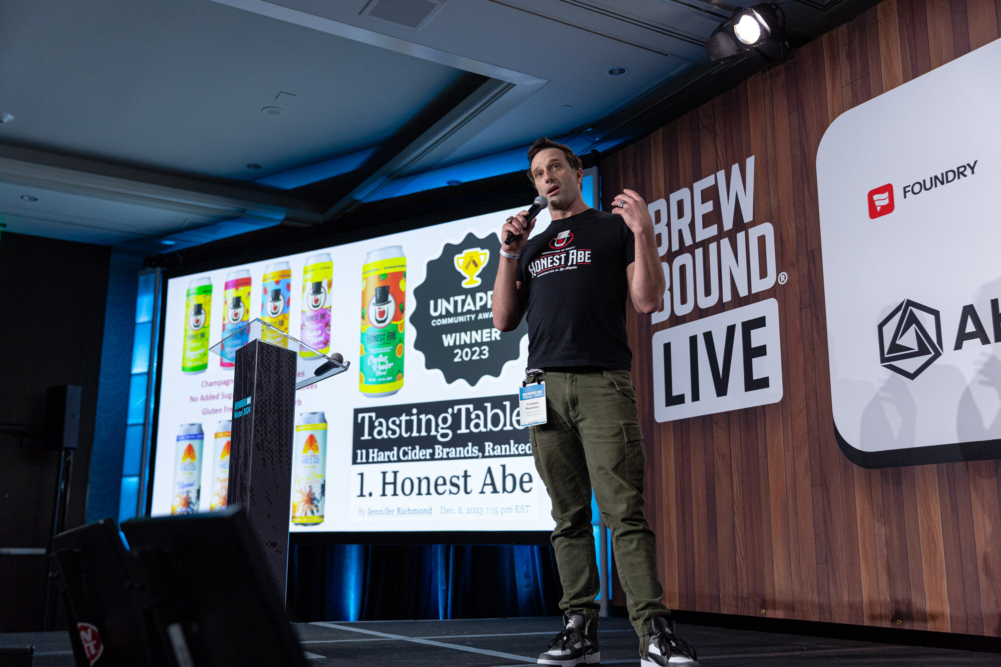 A man stands on stage next to a screen showing Untappd Community Awards and Tasting Table cider rankings.