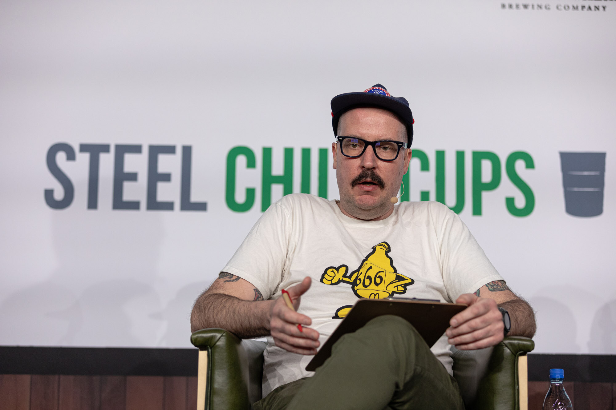A man in a navy cap and glasses with a mustache sits in front of a Steel Chill-Cups sign holding a clipboard.