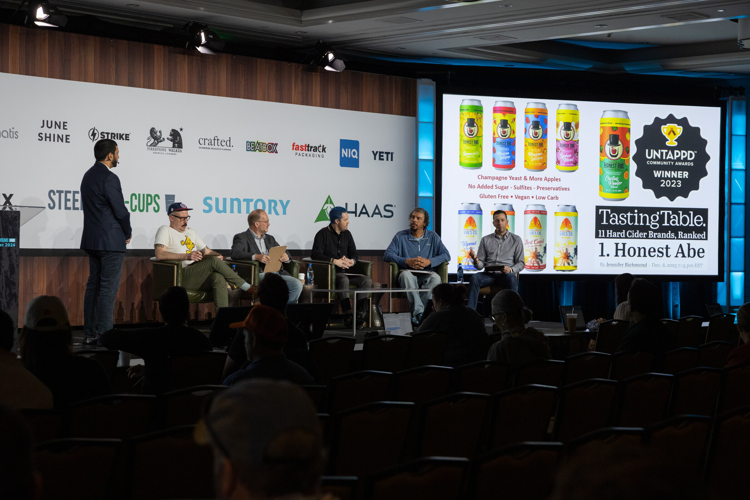 A man stands at left of a stage where five judges sit, with an Untappd and Tasting Table slide on screen.