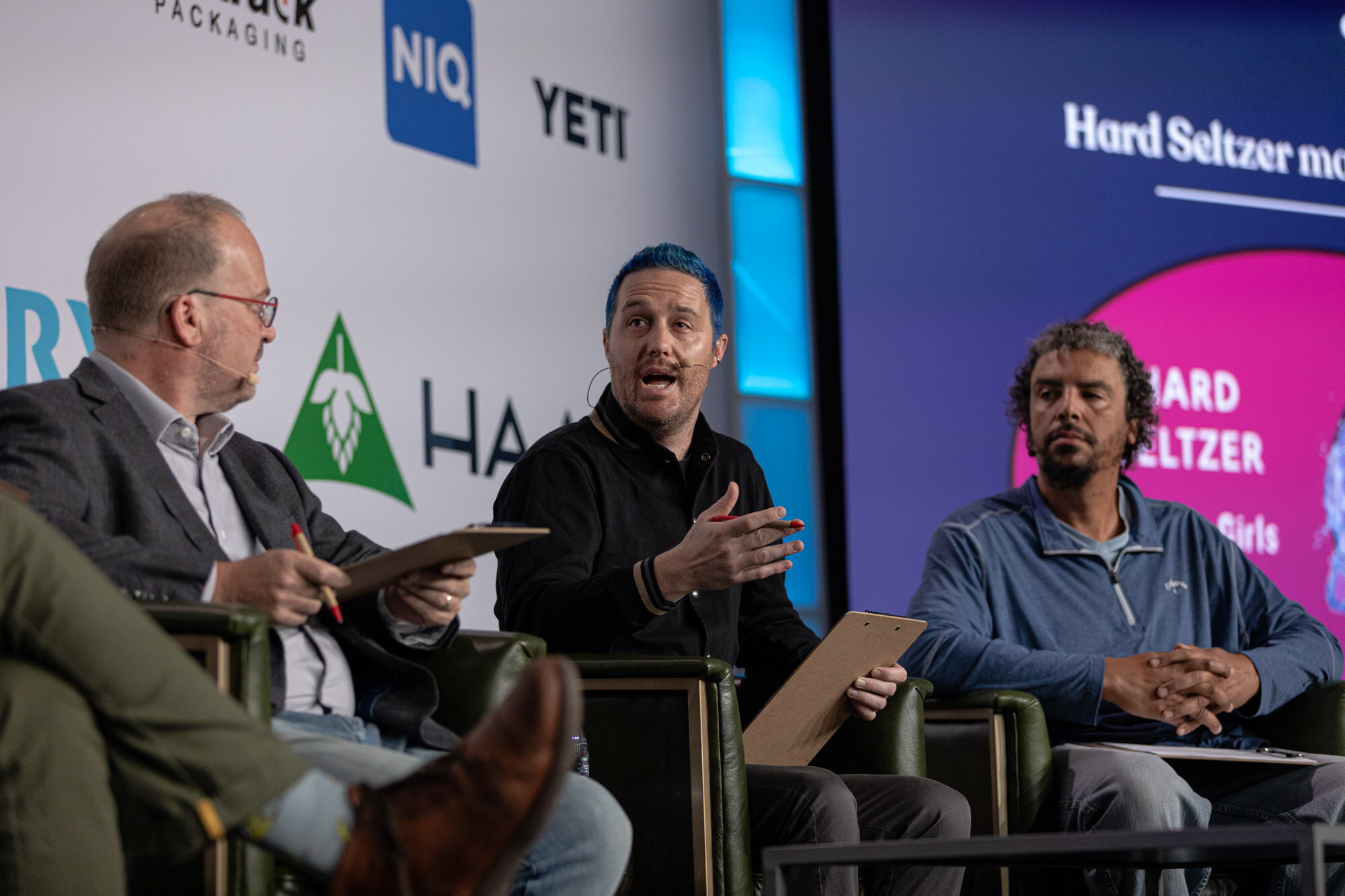 Three men sit in armchairs with a hard seltzer slide on the screen behind them.