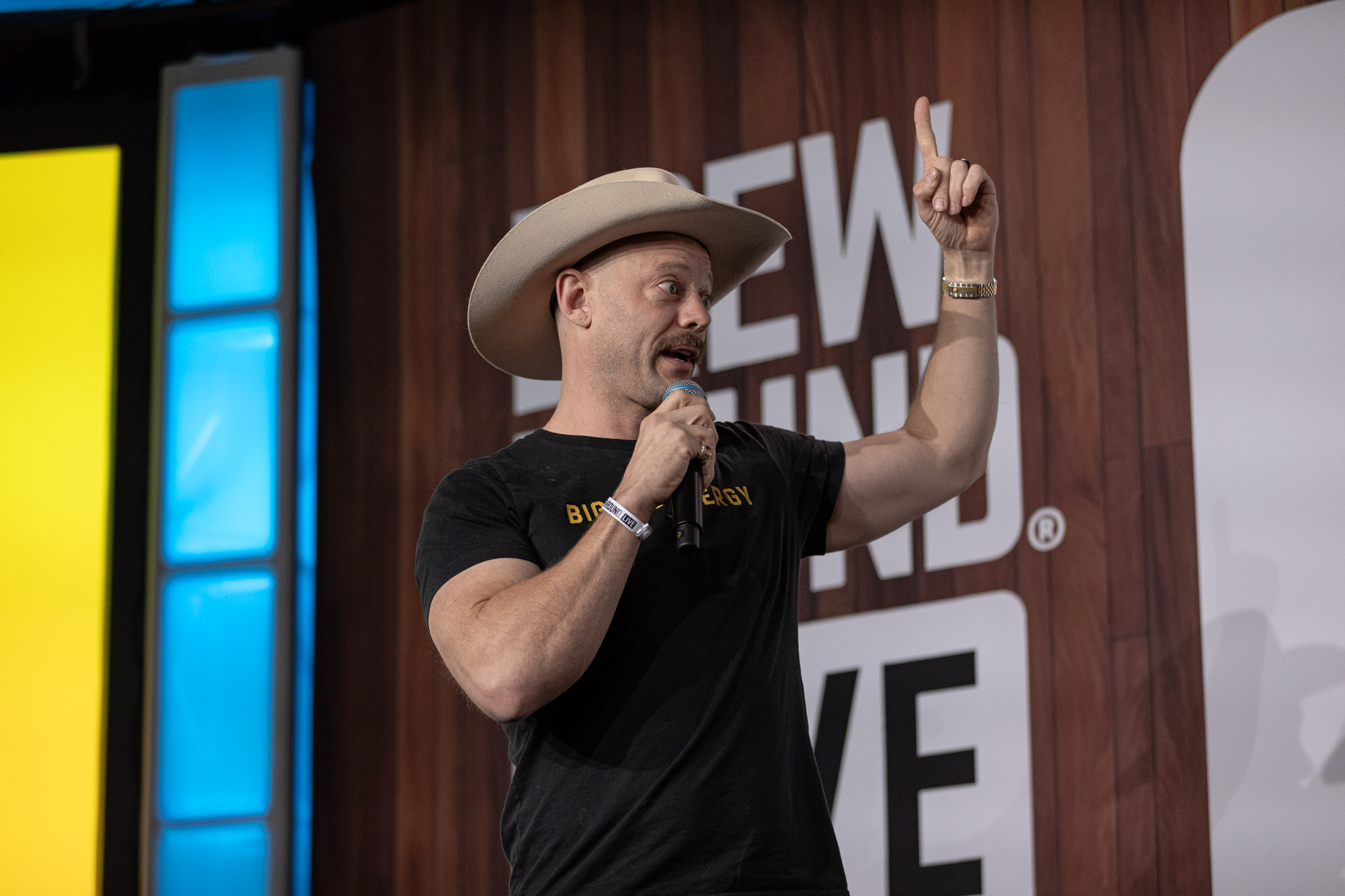 A man in a cowboy hat and a Big Dad Energy shirt raises one finger in front of a yellow slide.
