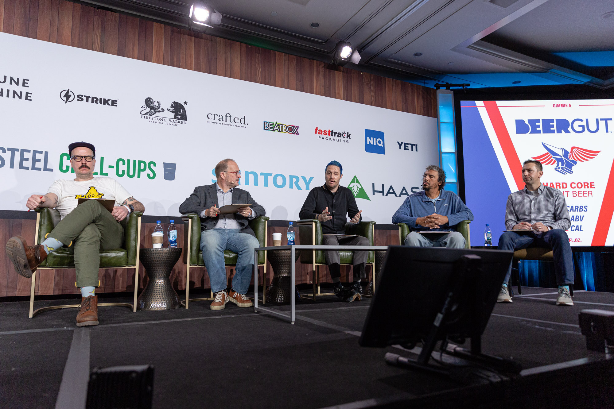 Five men sit in armchairs on stage next to a screen with a blue and red Beergut slide.