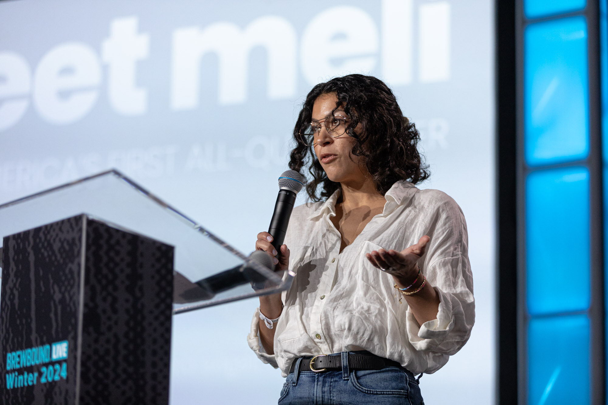 A woman in a white blouse speaks into a microphone in front of a meli slide.