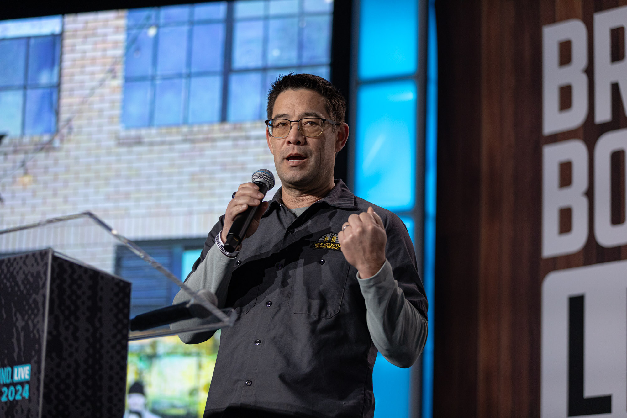 A man with glasses in a gray work shirt clenches a fist while speaking in front of a brick building slide.