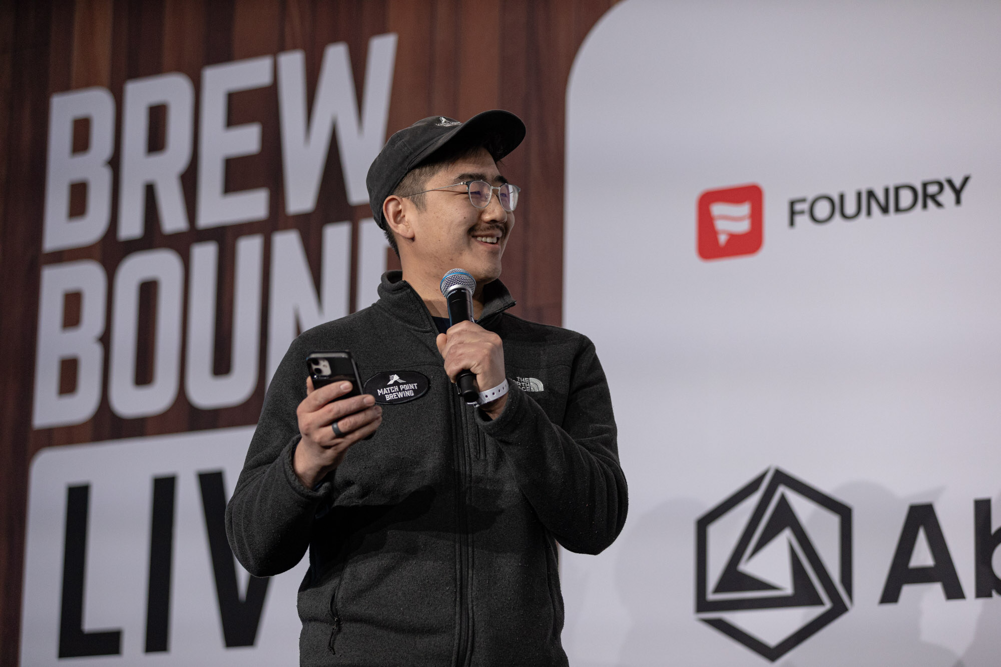 A man in a black cap and fleece holds a phone and microphone in front of a Foundry sponsor panel.