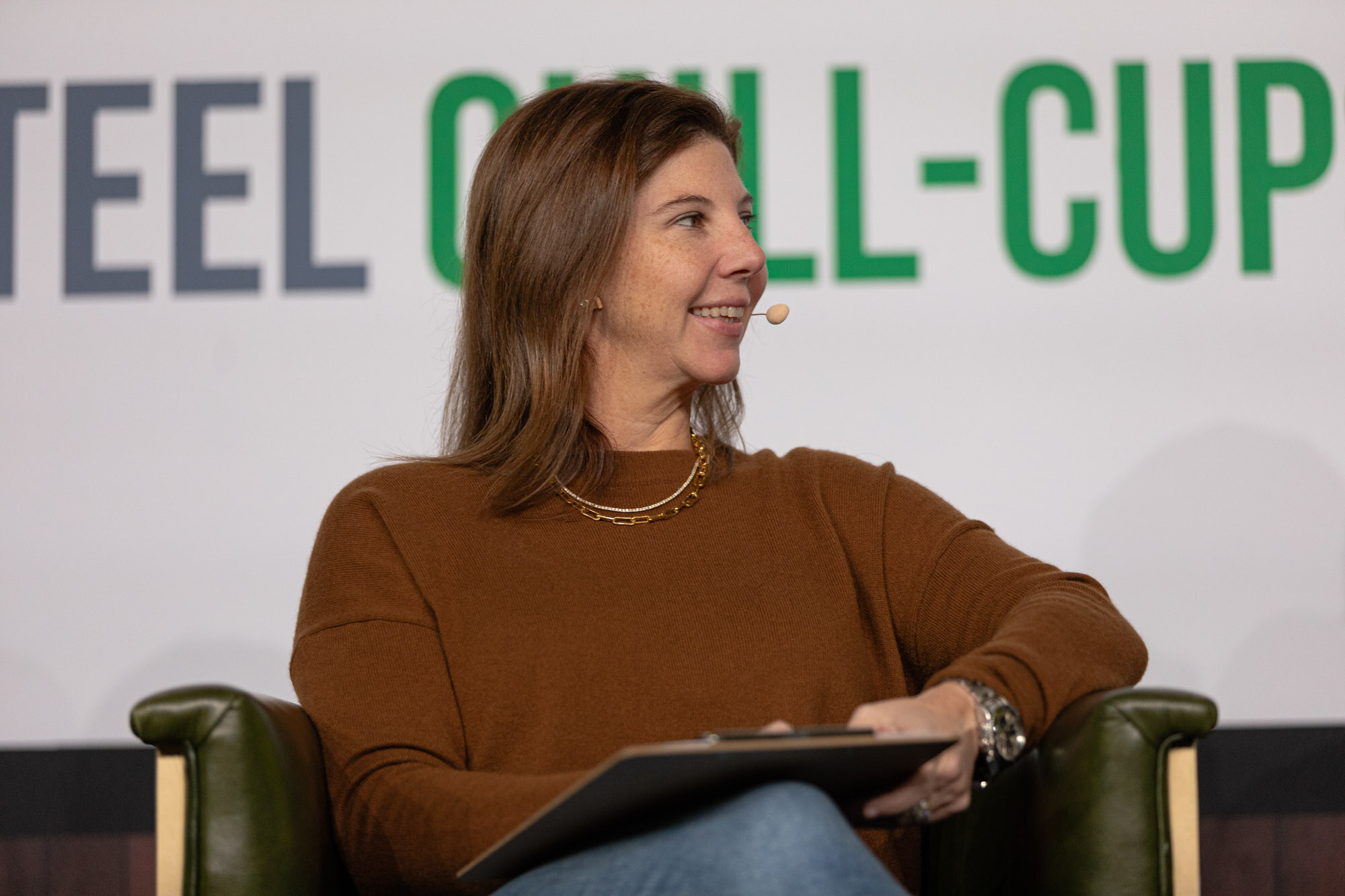 A woman in a brown sweater holds a clipboard while seated in front of a Steel Chill-Cups sign.
