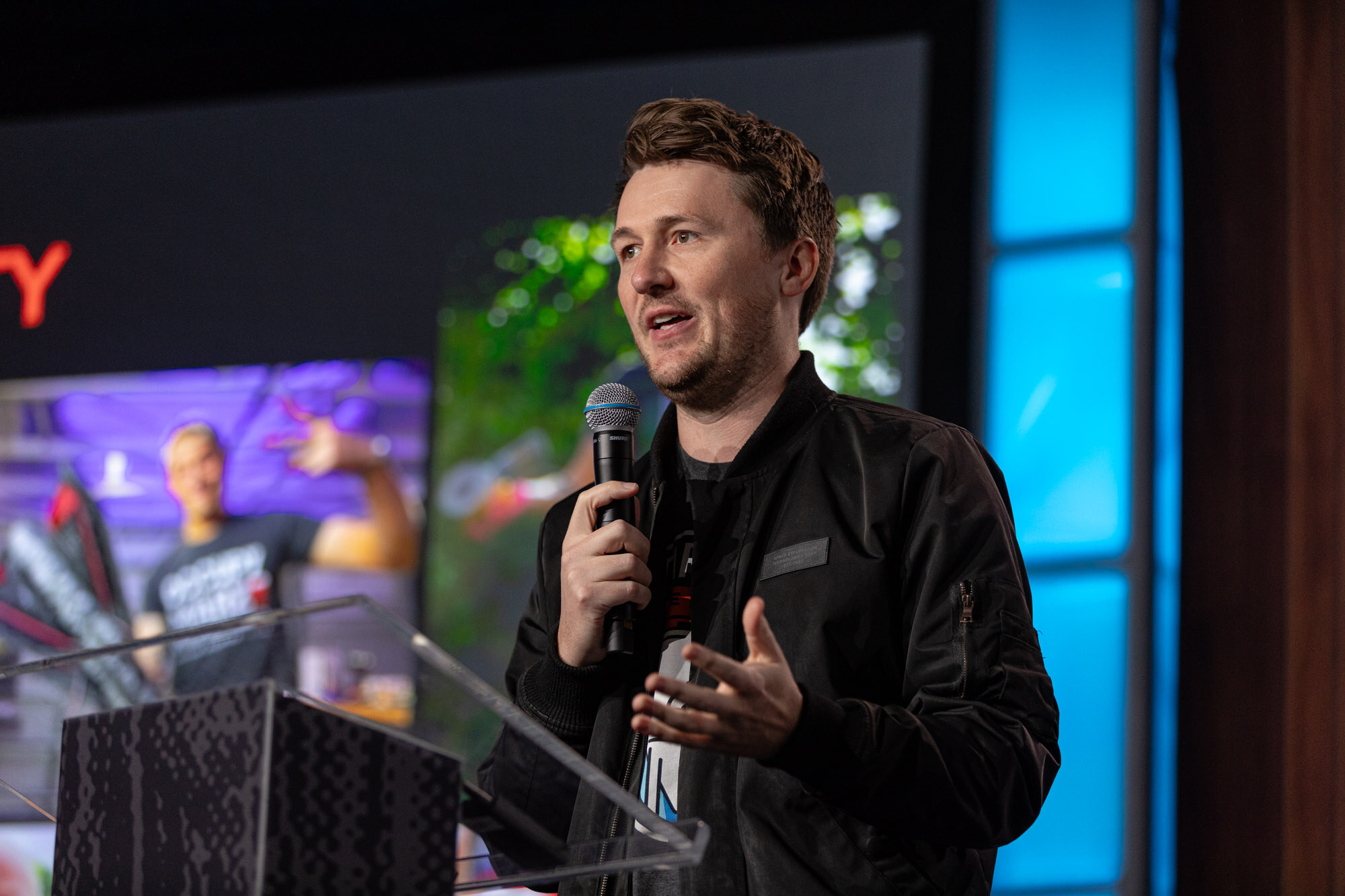 A man in a black jacket speaks into a microphone at a podium on stage.