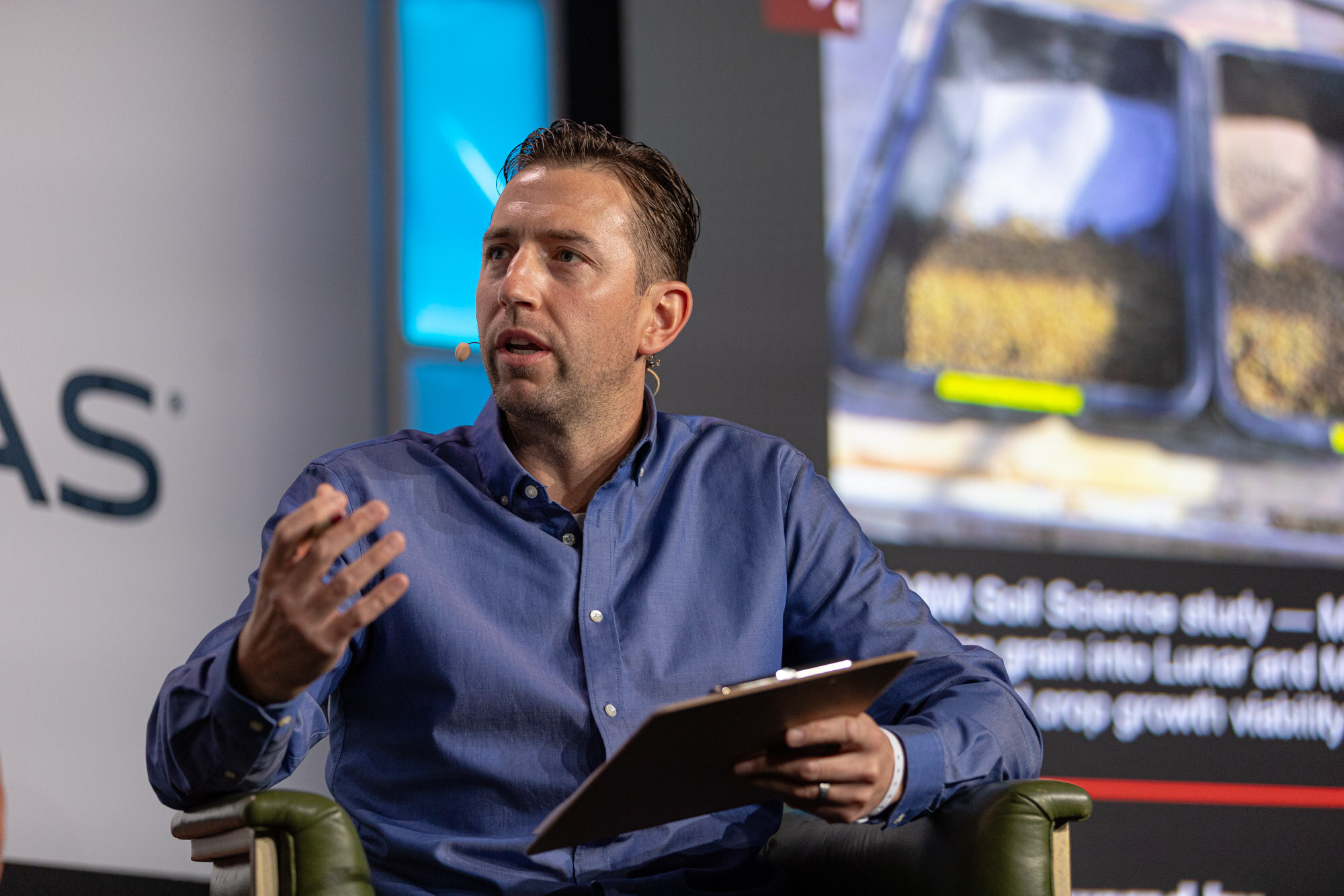 A man in a blue shirt holds a clipboard while seated beside a screen.