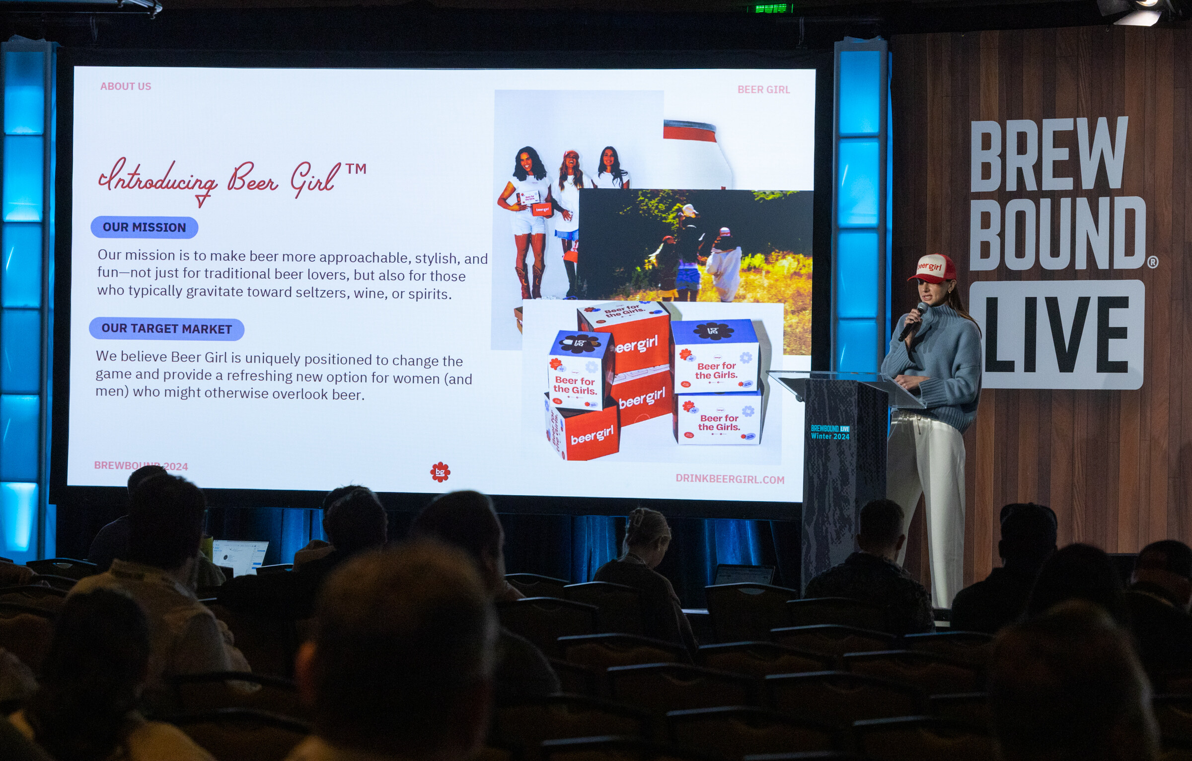 A woman in a cap stands at a podium beside a large screen showing an Introducing Beer Girl slide.