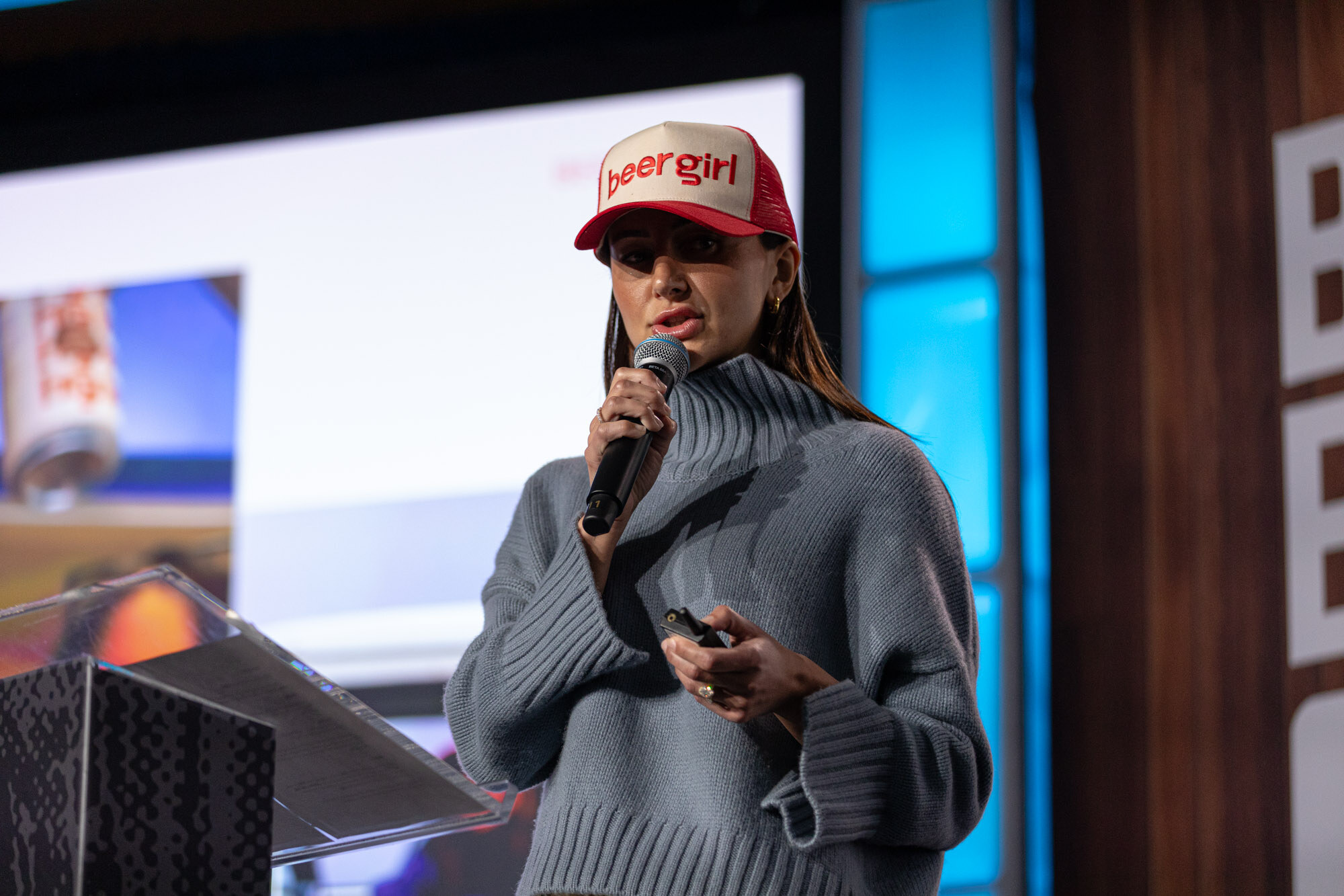 A woman in a gray sweater and a red and white cap with beergirl written on it speaks at a podium.