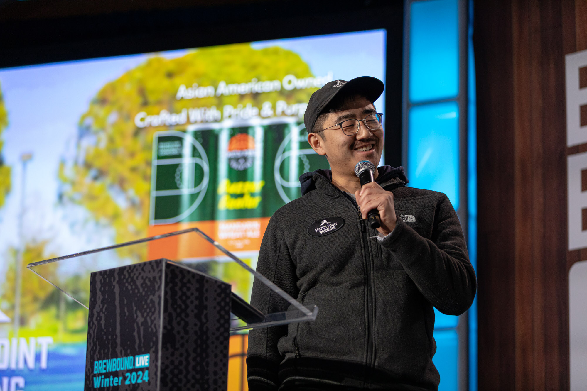 A man in a cap and fleece smiles at a podium in front of a slide for Match Point Brewing.