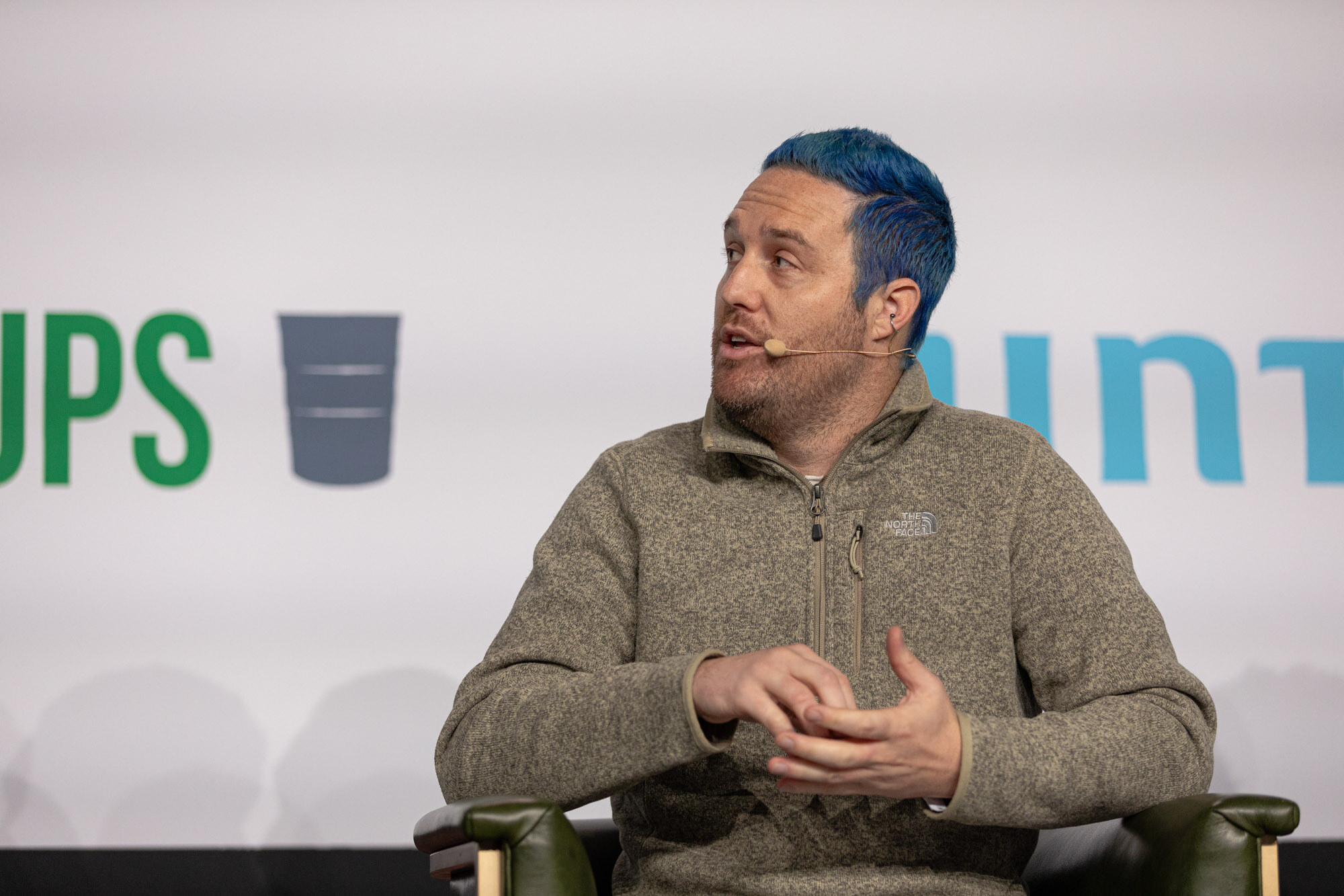 A man with blue hair in a quarter-zip sweater sits in an armchair and gestures.
