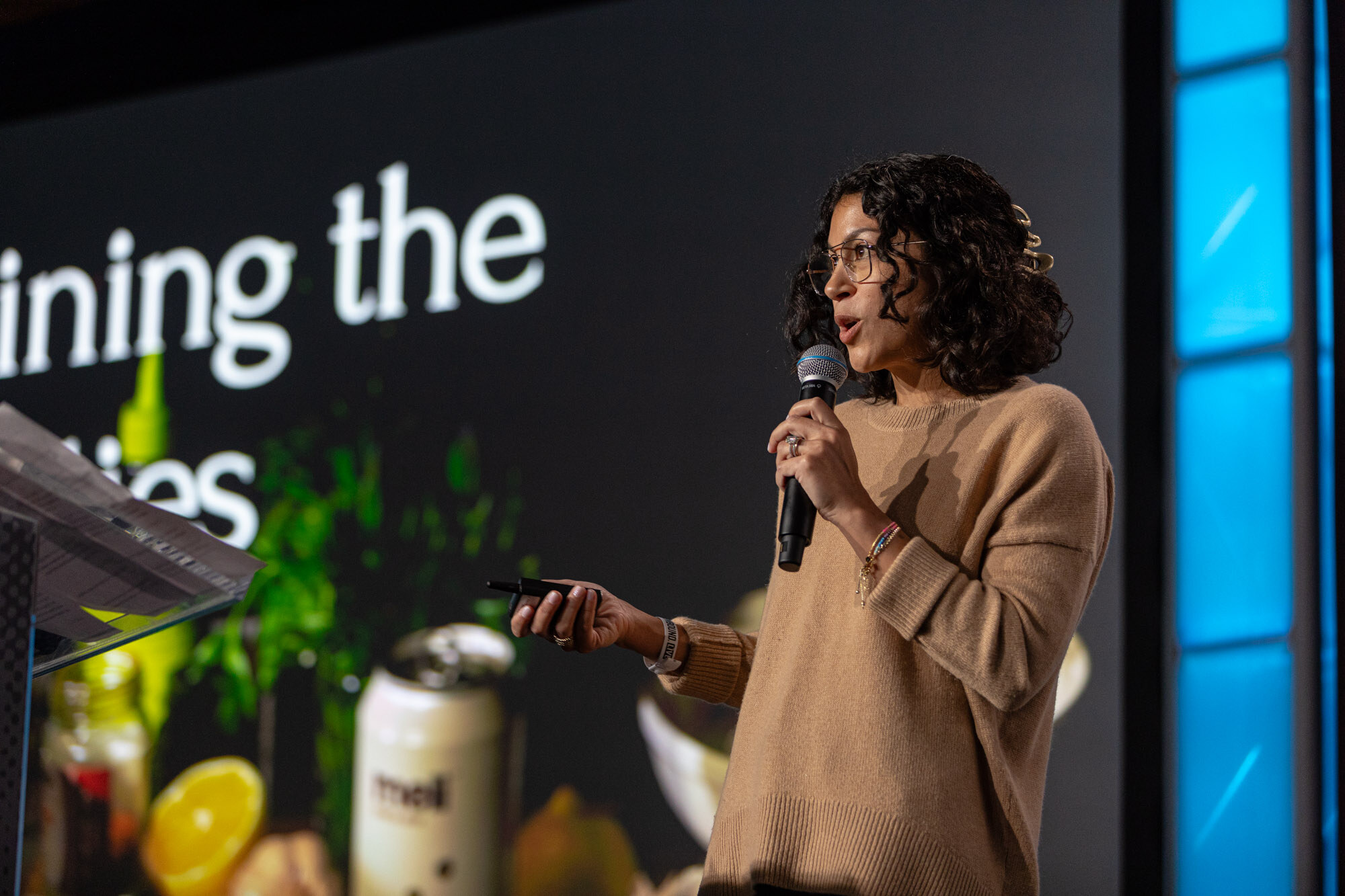 A woman in a tan sweater holds a microphone and a clicker beside a slide with a can of meli.
