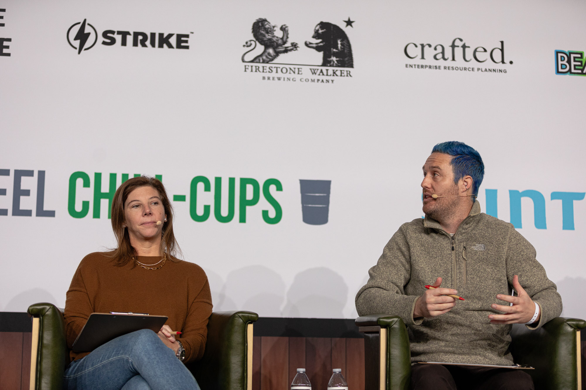 A woman in a brown sweater and a man with blue hair sit side by side in front of a sponsor wall.