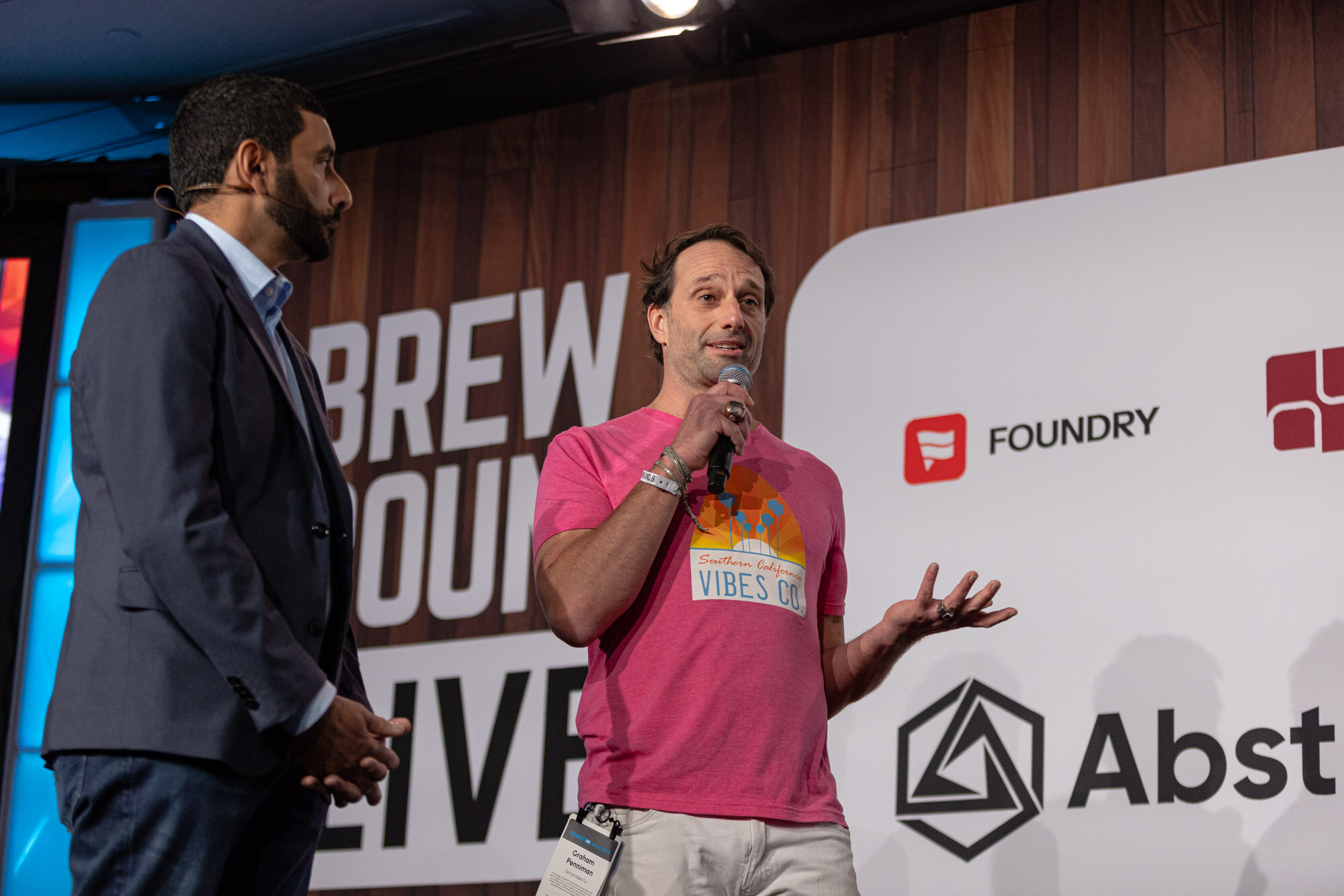 A man in a suit stands beside a man in a pink shirt on stage in front of a Brewbound Live backdrop.