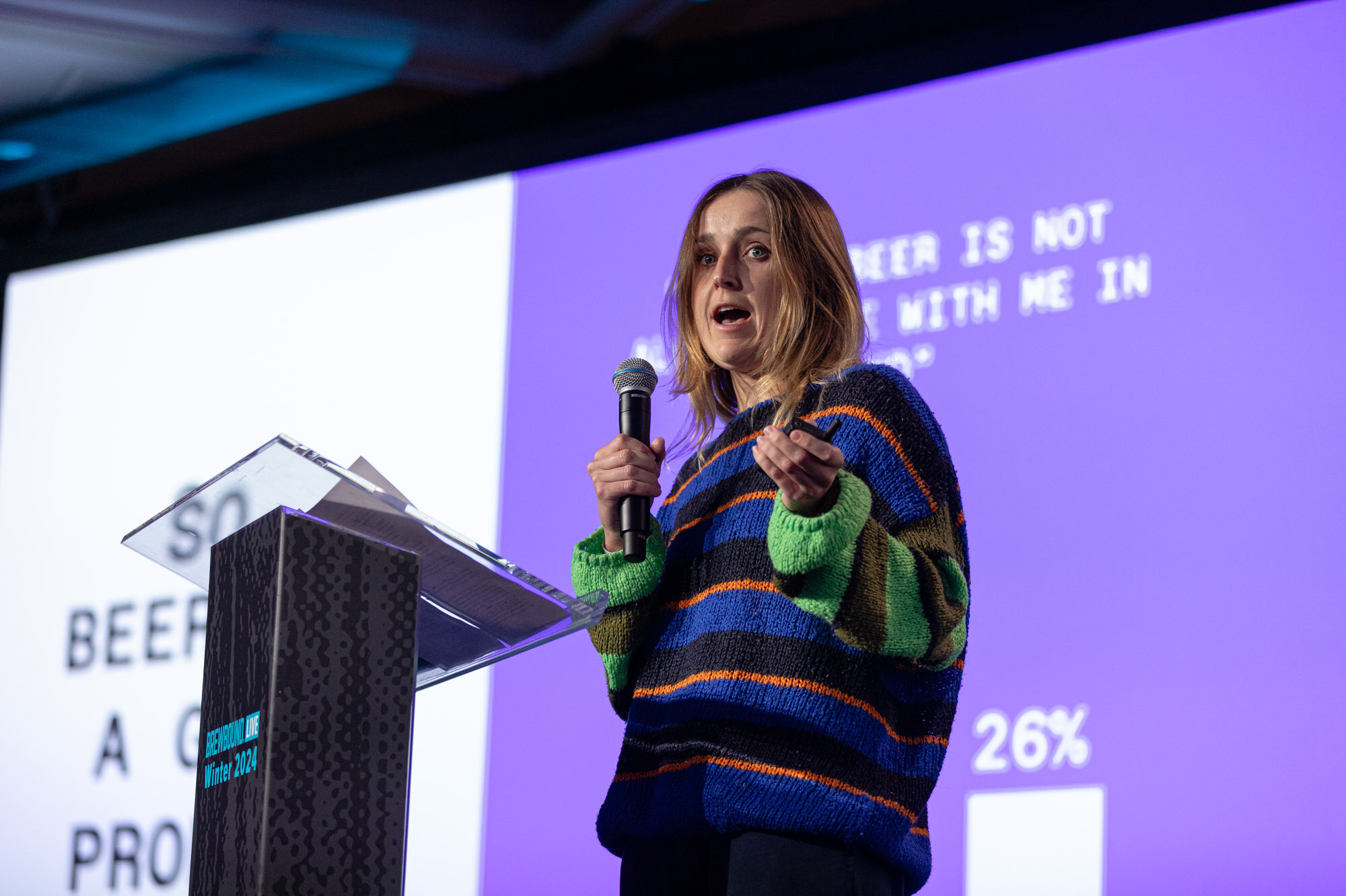 A woman in a colorful striped sweater speaks at a podium in front of a purple slide.