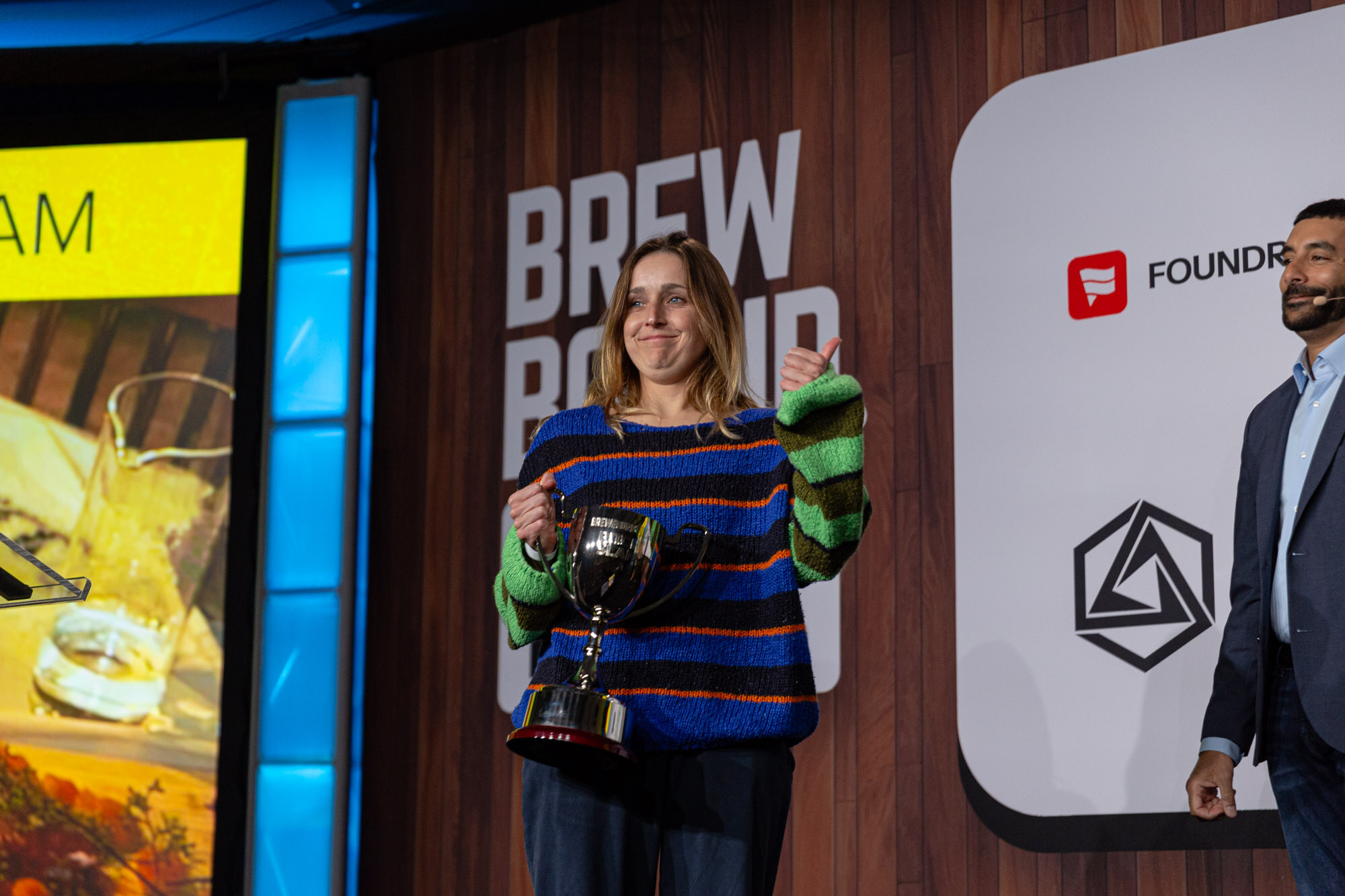 A woman in a colorful striped sweater holds a trophy and gives a thumbs up on stage.