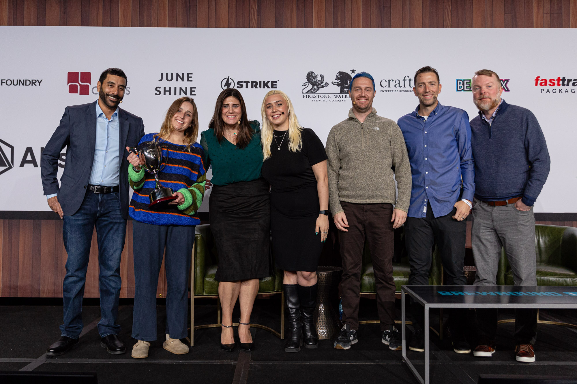 Seven people stand in a line on stage, one holding a trophy, in front of the sponsor wall.