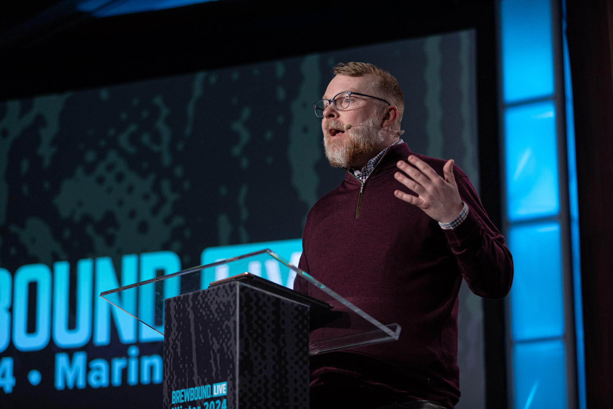A bearded man in a maroon sweater speaks from a podium on stage in front of a large screen.