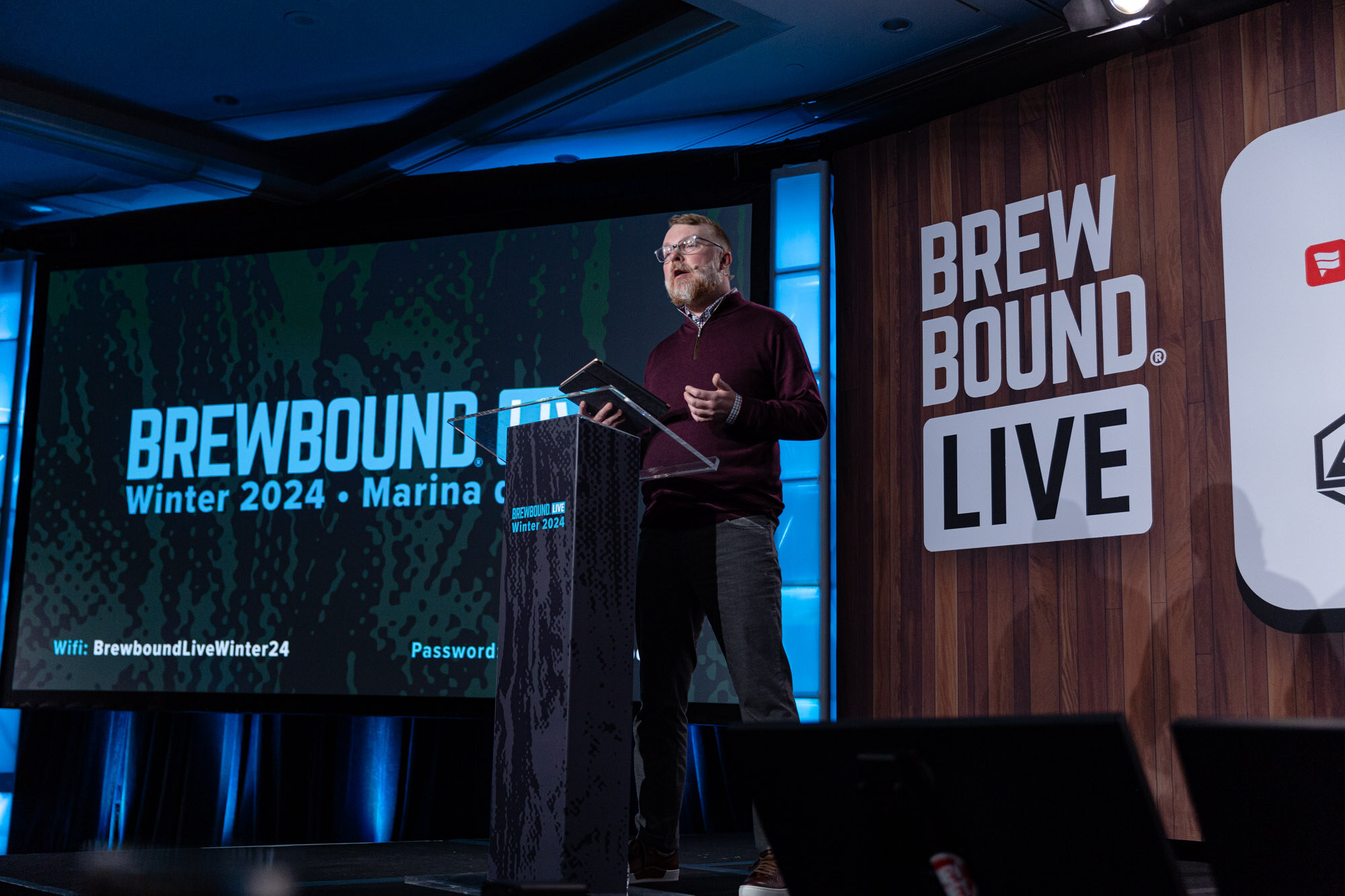 A man stands at a podium on stage beside a large screen reading Brewbound Live Winter 2024, Marina del Rey, with a Brewbound Live backdrop behind him.