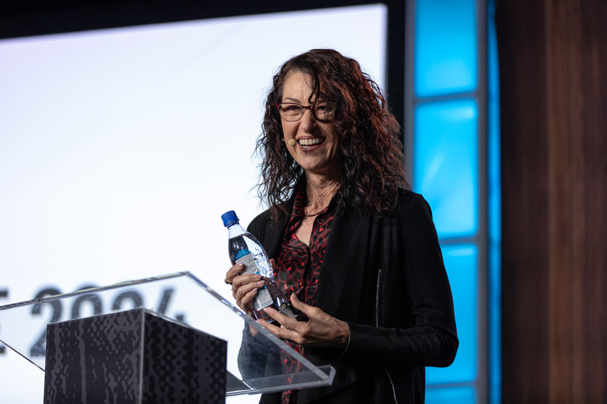 A woman with long curly hair smiles while holding a water bottle at a podium.