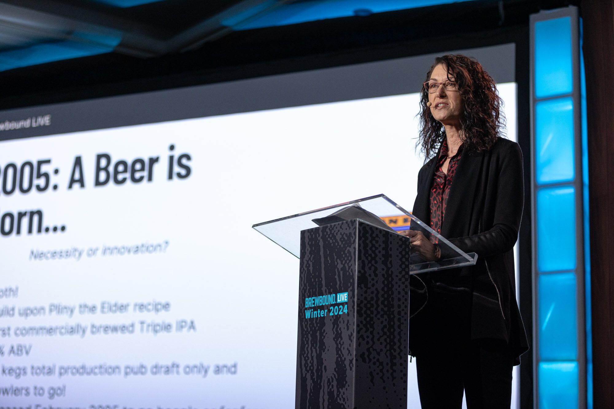 A woman stands at a clear podium beside a screen with a slide titled 2005: A Beer is Born.